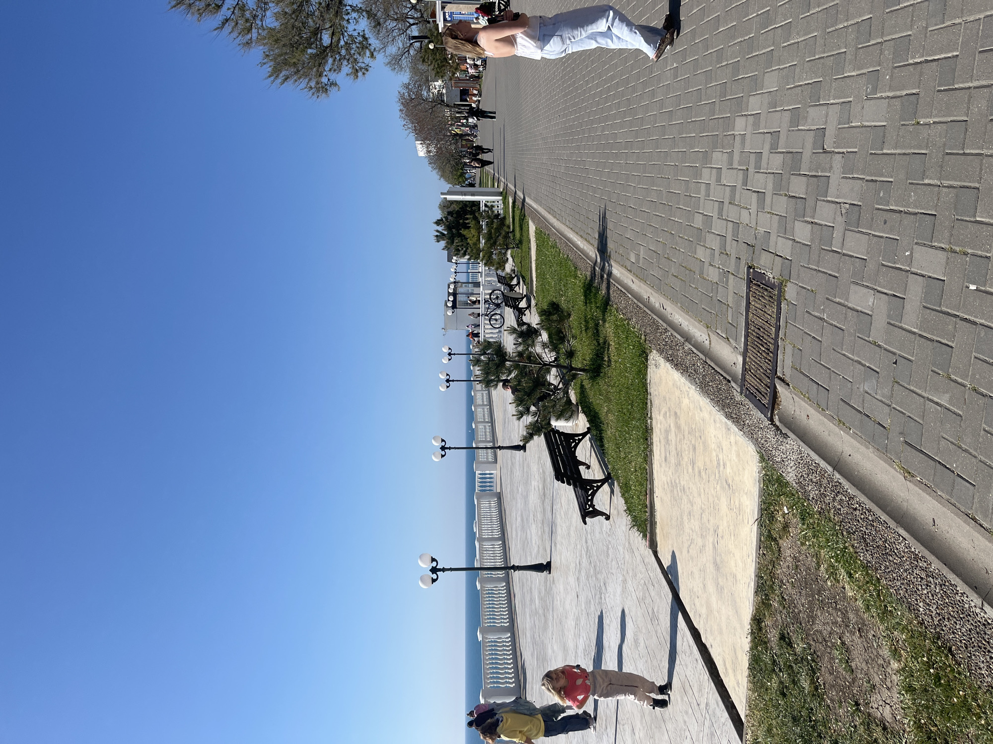 The image shows a promenade along the Black Sea in Anapa, Russia on a sunny day. The sky is clear and blue. The promenade is paved with gray rectangular stones. There are trees, grass, and benches along the edge. Street lights line the promenade. People are walking on the promenade. A group of three people is standing near the railing, and one person is walking further down the path.