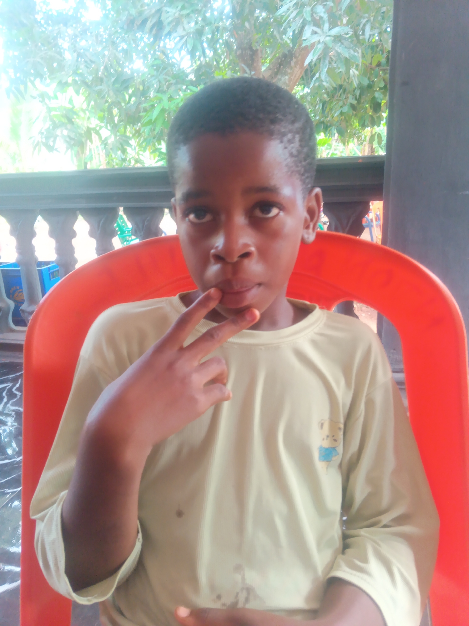 A young Black child is depicted in a portrait-style photograph, seated in a vibrant orange plastic chair. The child is looking directly at the camera with a neutral, calm expression. They wear a light yellow or pale green long-sleeved t-shirt, which features a small cartoon graphic of a bear wearing a blue shirt on the left side of the chest. The child's left hand is raised towards their face, with the index and middle fingers extended in a peace or victory sign, while the thumb rests near their lips.
The setting appears to be an outdoor or semi-outdoor area, consistent with a location in Eziudo, Nigeria. Behind the child, a light-colored balustrade or railing with decorative pillars runs horizontally across the mid-ground. Beyond the railing, abundant green foliage and tree branches are visible, suggesting lush vegetation and an open-air environment. A dark vertical pillar or wall segment is noticeable on the right side of the frame. The scene is brightly lit by natural light, indicating it is daytime. No discernible text is present in the image apart from the bear graphic.