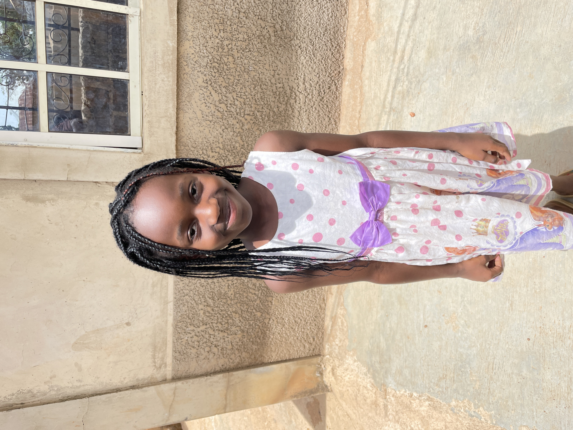A young girl stands outdoors near a building with a window, possibly in Jos, Nigeria. She wears a light-colored dress with pink polka dots and a purple bow. The building's exterior is a textured beige. The ground is paved.
