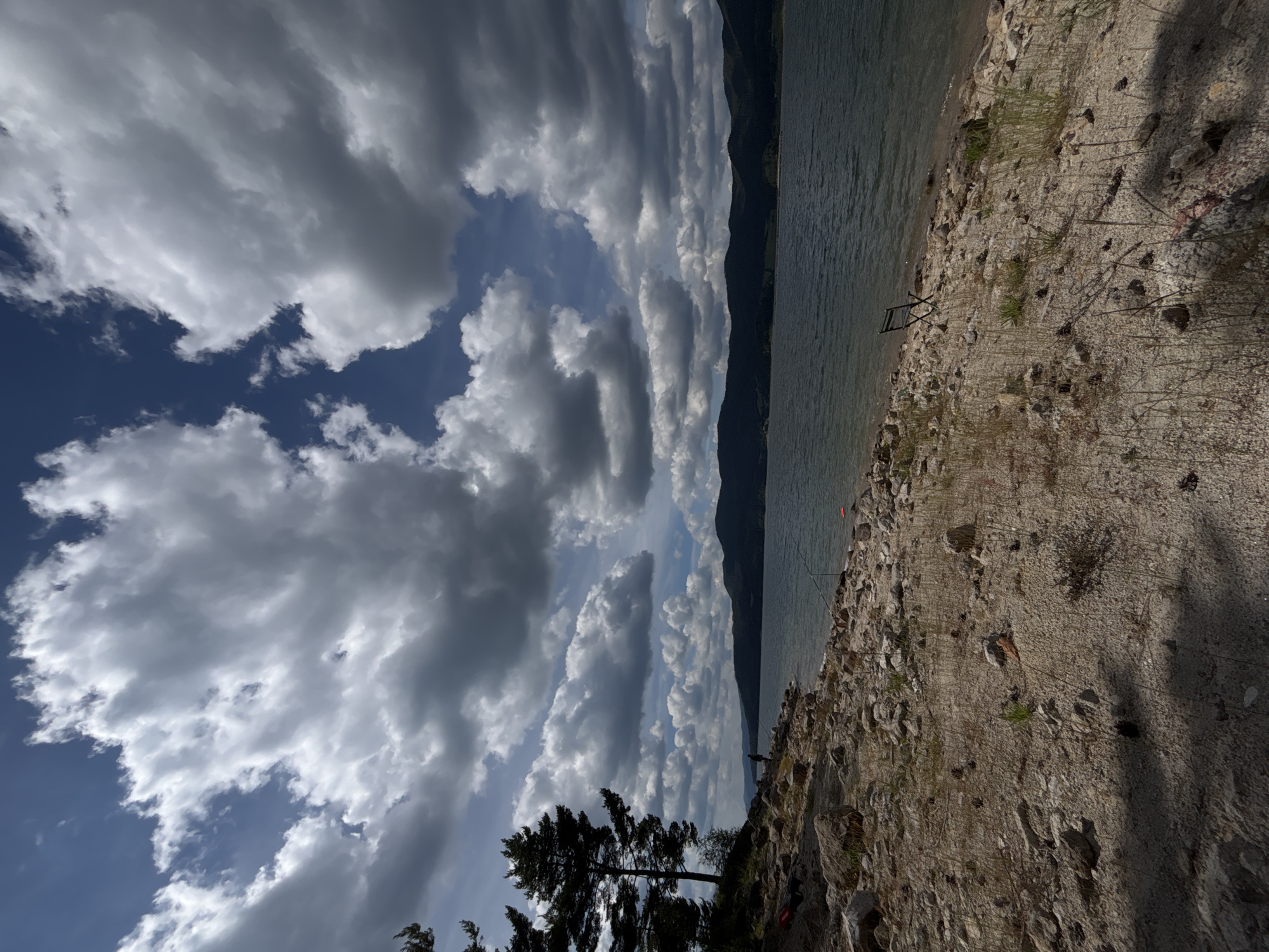 The image depicts a rocky shoreline on a body of water, likely a lake or reservoir, with mountains in the background. The sky is filled with dramatic, grey and white cumulus clouds, suggesting a dynamic weather system, possibly indicative of afternoon. The ground is covered in small to medium-sized rocks and sparse, dry vegetation.
In the middle distance, a dark, silhouetted tree stands against the lighter sky. On the shore, a few fishing rods are visible, one with a person standing nearby, suggesting a fishing activity. The water appears calm, reflecting the sky and surrounding landscape. The overall mood is serene and natural, with a sense of solitude. The lighting suggests it is daytime, with shadows indicating the sun's position, though no specific time can be precisely determined. No text is visible in the image.