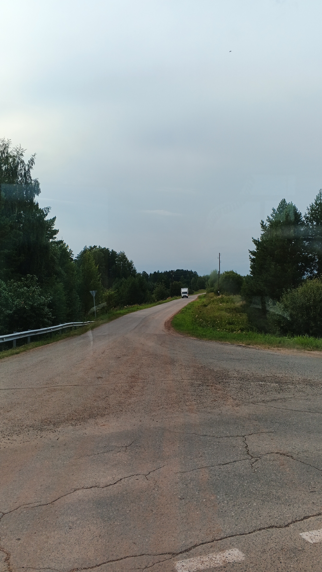 The image depicts a rural road scene in Киясово, Russia. A paved road, showing signs of wear with cracks, curves through a landscape of dense green trees on either side. A white guardrail lines the left side of the road. In the distance, a white truck is visible driving away from the camera. The sky is overcast with a pale, diffused light. The scene suggests a quiet, possibly remote, setting within the region.
