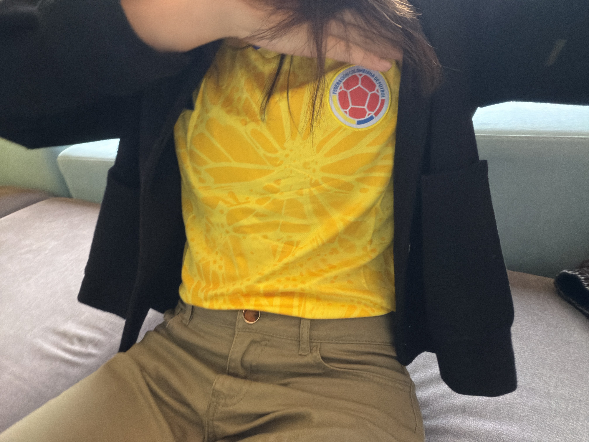 The image features a person captured from the chest down, seated indoors on upholstered furniture. They are wearing a vibrant yellow t-shirt with a subtle, abstract pattern, layered with an open black jacket. Khaki-colored pants complete the attire, with a visible belt loop and a metallic button or ring near the waist. Dark hair cascades over one shoulder.
A prominent circular emblem is affixed to the right chest of the yellow t-shirt. It displays the text "FEDERACIÓN COLOMBIANA DE FÚTBOL" along its upper arc. The emblem's center depicts a stylized football, incorporating the red, yellow, and blue colors of the Colombian flag, strongly indicating a connection to Colombian sports. This detail is consistent with the provided location context of Bogota, Colombia. The person's posture suggests a casual moment of rest, with one hand slightly raised and their face out of frame. The indoor setting is softly lit, with blurred elements of furniture and potentially a window in the background.
