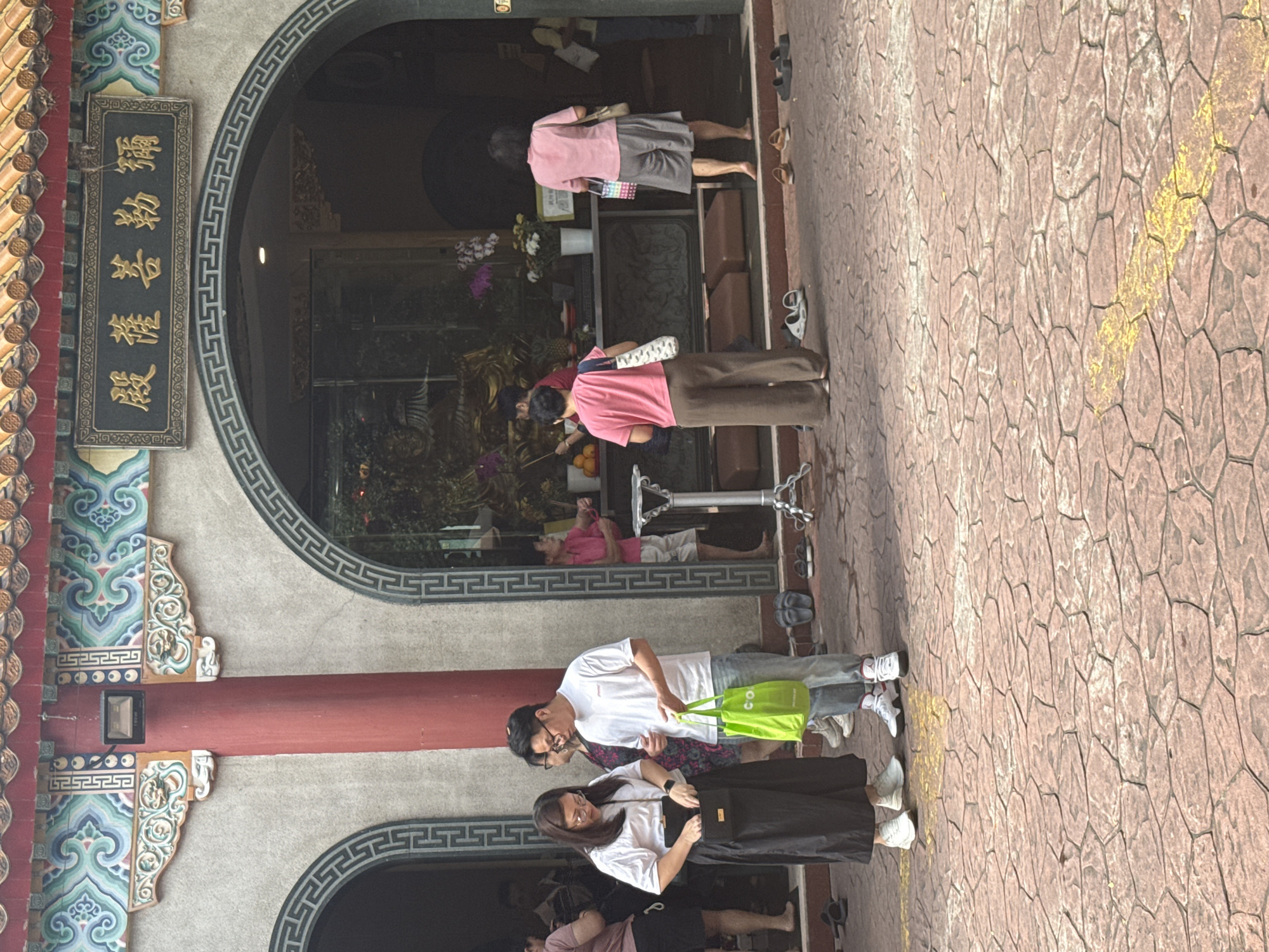 The image shows a group of people in what appears to be a temple in Kuala Lumpur, Malaysia. The architecture includes a traditional Chinese style with ornate decorations. There is a sign in Chinese script on the wall. People are standing around, some appearing to be praying or paying respects inside the temple. There are several entrances with decorative archways. The ground is paved with patterned stones. A couple stand near the lower entrance.