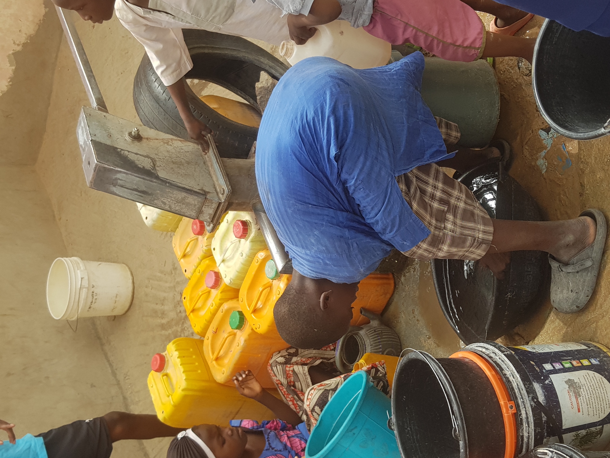 People are collecting water in what appears to be an urban environment in Dutsinma A, Nigeria. Individuals fill various containers, including plastic jerrycans (predominantly yellow) and buckets, using a pump. The area around the pump is muddy, with several containers scattered nearby. One person is actively filling a black bucket while another appears to be operating the pump. A tire is present near the pump mechanism. The walls of a building form part of the backdrop.