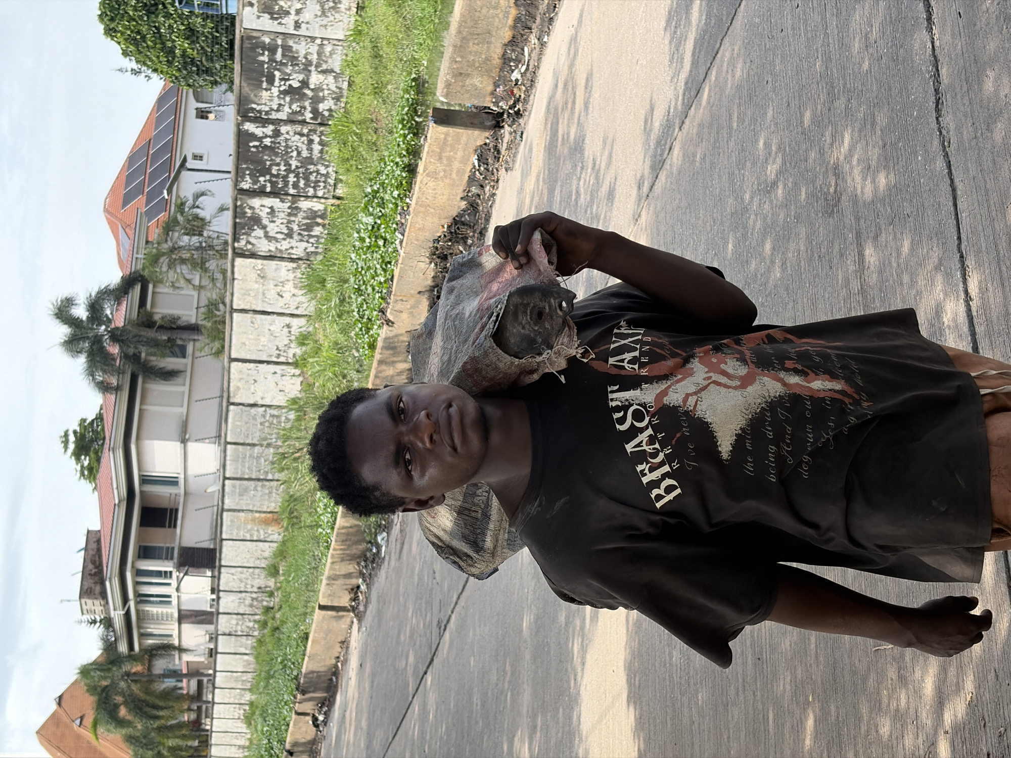 A young Black male stands on a paved road, facing the camera with a weary expression. He is carrying a large, dirty, burlap sack over his right shoulder, its contents vaguely visible as a dark, lumpy mass. The man wears a dark t-shirt with white text that reads "BRAST RETRO REND" and other illegible words. He also wears tan shorts. The background features a narrow strip of overgrown green grass and a concrete barrier. Beyond the grass, a multi-story building with a red-tiled roof and solar panels is visible, along with palm trees. The scene appears to be outdoors during the daytime, with dappled sunlight casting shadows on the road. The overall impression is one of a person engaged in difficult labor or carrying a heavy burden.