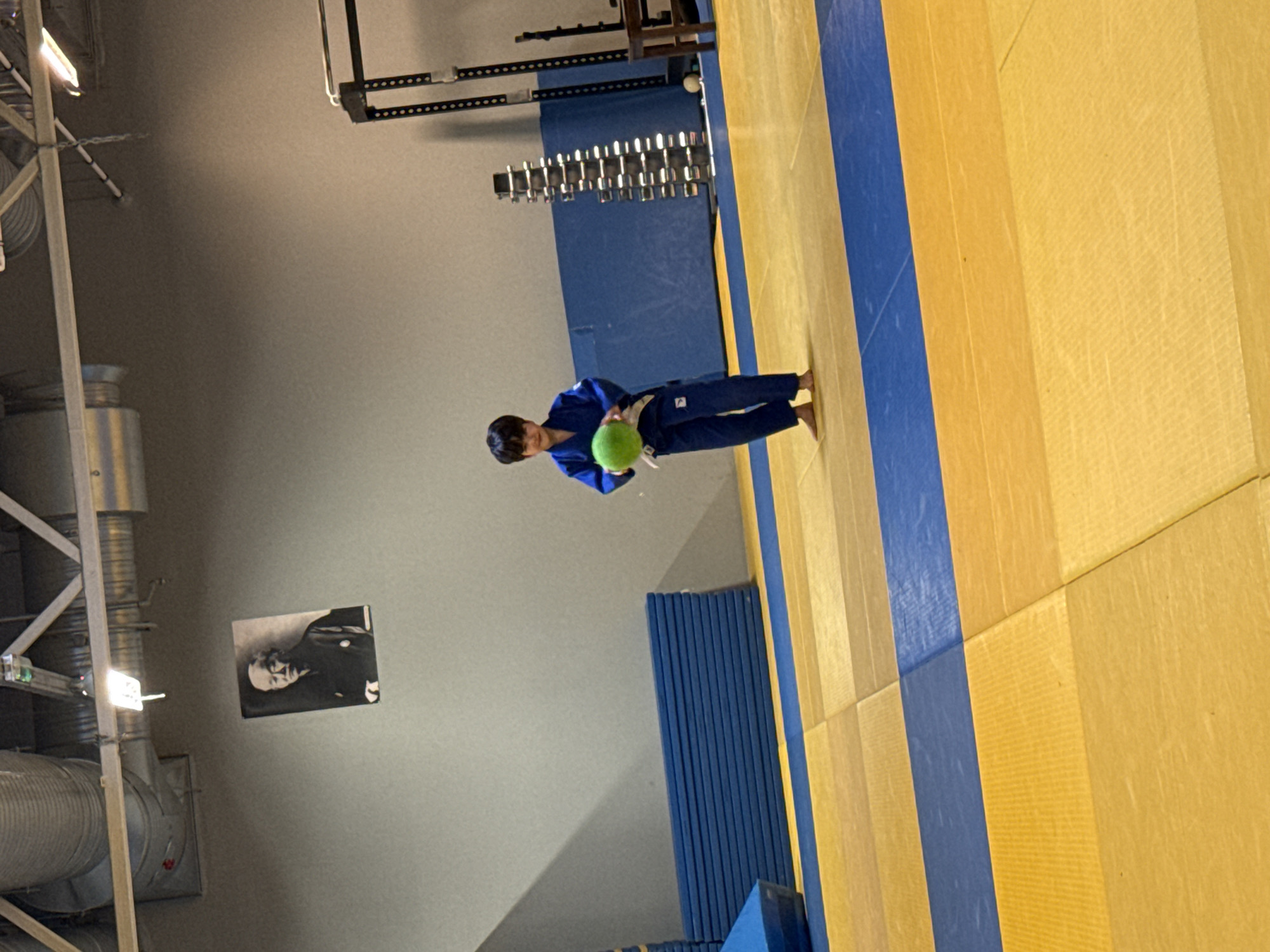 A young boy dressed in a blue judo gi and white belt stands barefoot on a yellow and blue mat in what appears to be a gymnasium or dojo. He is holding a green ball in his hands, looking towards the camera with a slight smile. In the background, a black and white portrait of an older man hangs on a grey wall. Further back, blue mats are stacked, and weightlifting equipment, including a rack of dumbbells, is visible. The ceiling features industrial lighting and ventilation systems. The floor is composed of interlocking mats, predominantly yellow with blue stripes.