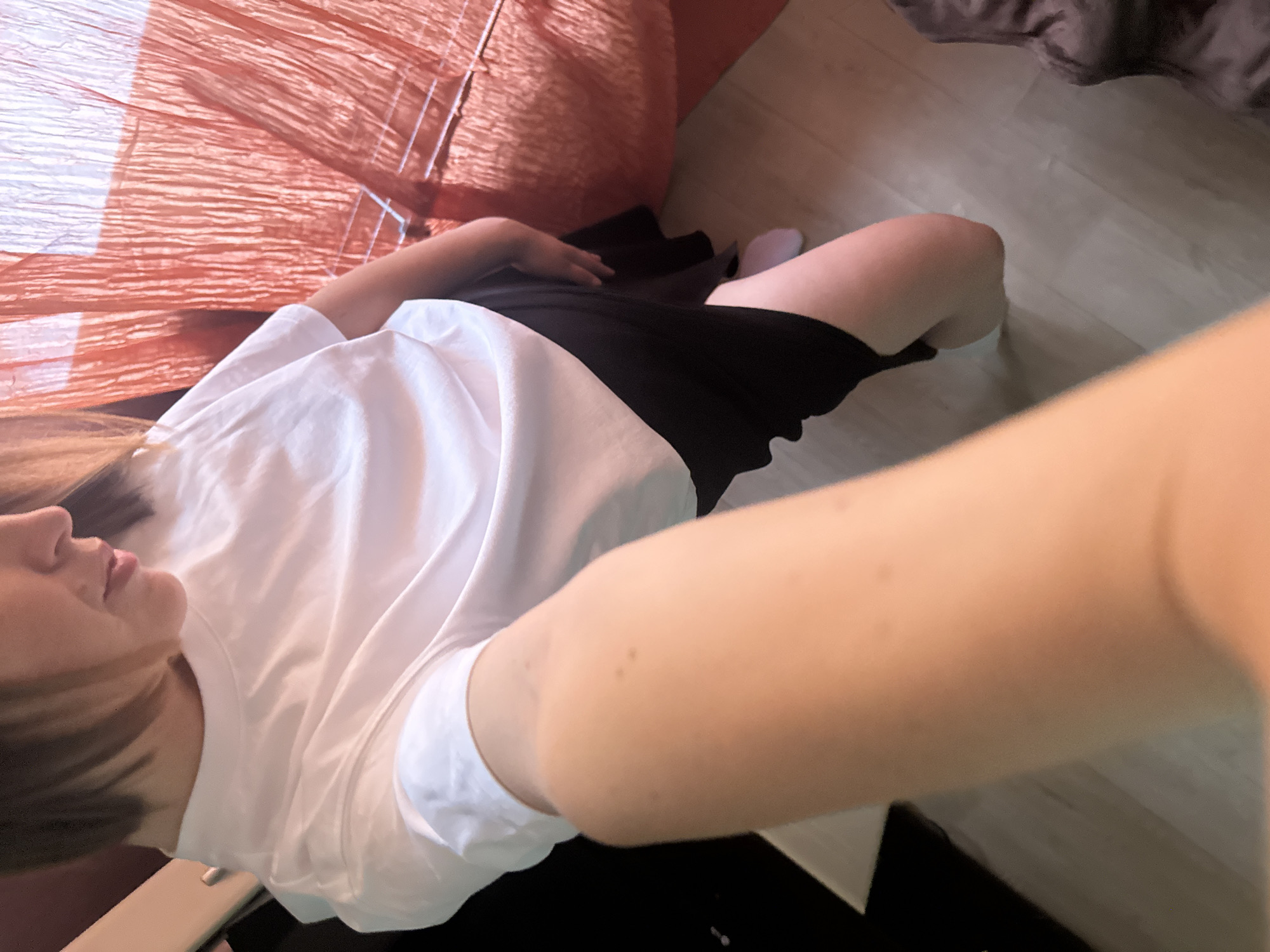 A first-person, high-angle shot depicts a woman lying on what appears to be a bed or soft surface with an orange textured covering. She is wearing a white t-shirt and a dark-colored garment, possibly a skirt or shorts. A portion of her leg and a sock are visible on a light-colored wooden floor. Part of a pillow is visible in the upper right of the image. The image does not provide any details to determine the location of the scene in the city of Chaikovsky, Russia. The woman seems to be taking a selfie, as her arm extends into the foreground.