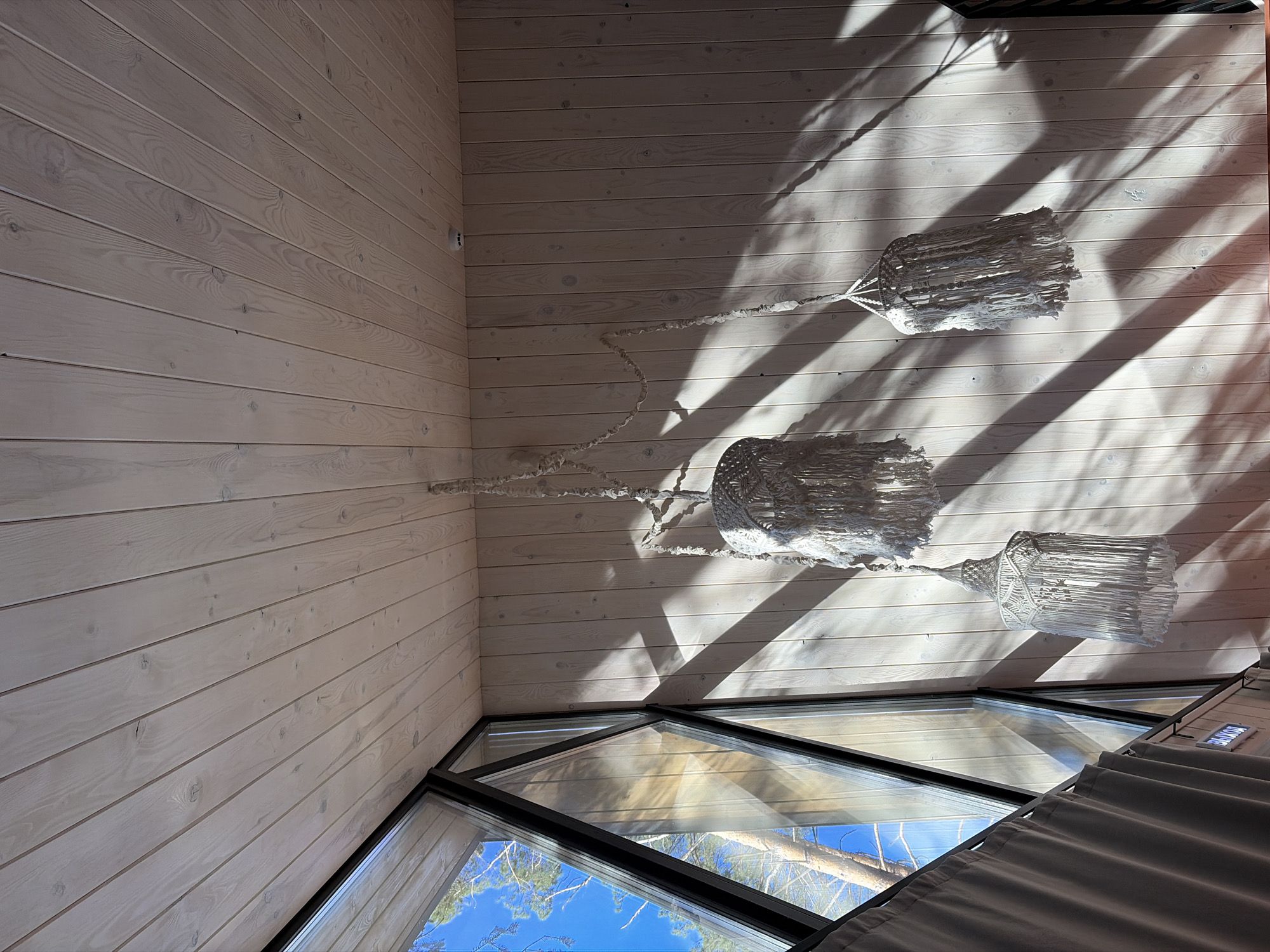 Captured from a low-angle perspective, the photograph showcases the interior of a structure in Мишкино, Russia. The walls and ceiling are clad in light-colored wood paneling. Natural light, filtered and patterned by external elements, casts distinct shadows across the ceiling. A multi-tiered macramé light fixture hangs centrally. Below, angular windows frame a view of the clear, blue sky and treetops. A section of neutral-toned curtains is visible on the right edge of the frame.