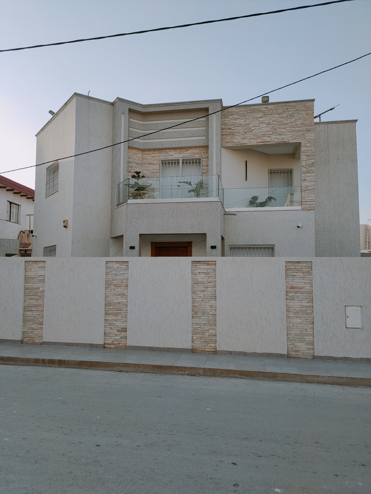 The image shows a modern, two-story house with a white, textured facade and stone accents. The house has a flat roof with some architectural detailing and features a balcony on the upper level enclosed by a glass railing. There are several windows with metal grates, and a brown wooden door is visible at the entrance. A light-colored concrete wall with vertical stone inlays surrounds the property, with a sidewalk and a street in front of it. The sky is clear and light blue, indicating it is daytime. The scene is located on a street in a city, with another building visible to the left, partially obscured by the main house. The overall impression is of a well-maintained residential property in a developed area.