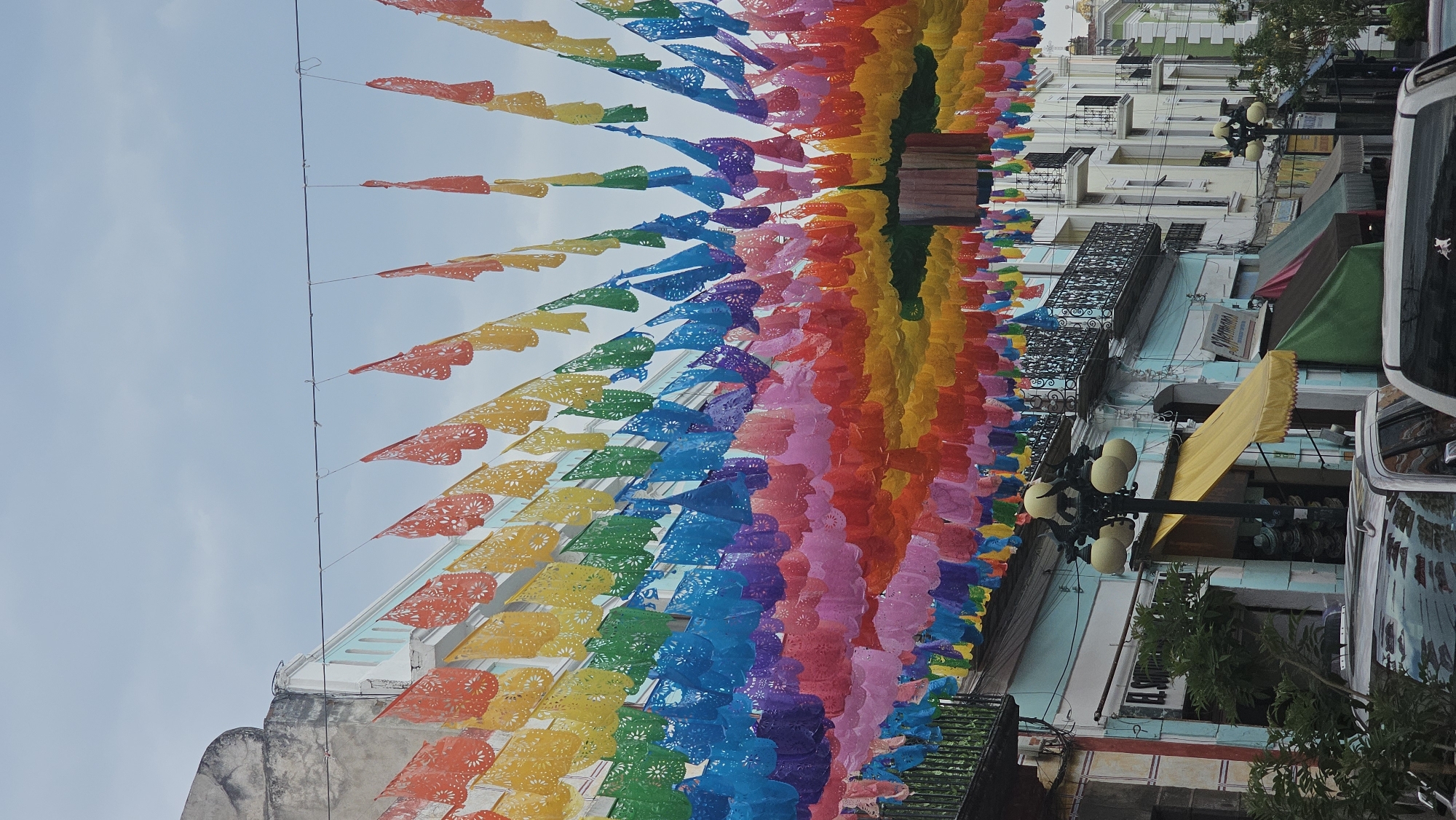 The image displays a vibrant street scene, likely in Puebla, Mexico, adorned with a spectacular overhead display of traditional *papel picado* banners. These colorful, perforated paper decorations are strung across the street in numerous lines, forming an arched canopy that spans the width of the thoroughfare. The *papel picado* exhibits a full rainbow spectrum of colors, including red, orange, yellow, green, blue, purple, and pink, creating a festive and cheerful atmosphere. Each banner is intricately cut with decorative patterns.
On the right side of the street, multi-story buildings with classic architectural features line the path. Some buildings have balconies with ornate wrought-iron railings and facades painted in white, light blue, and green. Below these structures, various storefronts are visible, shielded by awnings in yellow, green, and dark red. Street lamps with multiple globe-shaped lights are positioned along the sidewalk. Portions of parked vehicles, possibly cars or vans, are seen on the far right, suggesting a bustling urban setting. Scattered green foliage from trees or plants adds natural elements to the scene.
The bright, clear blue sky in the background indicates that the photograph was taken during the daytime. The presence of *papel picado* strongly suggests a celebration, festival, or a generally festive occasion. On a light blue building, a sign with partially visible text appears to read "MARIANA" or "MERCADO," and another sign below it might display "BAR." The overall mood conveyed by the image is one of joy, cultural richness, and vibrant activity.