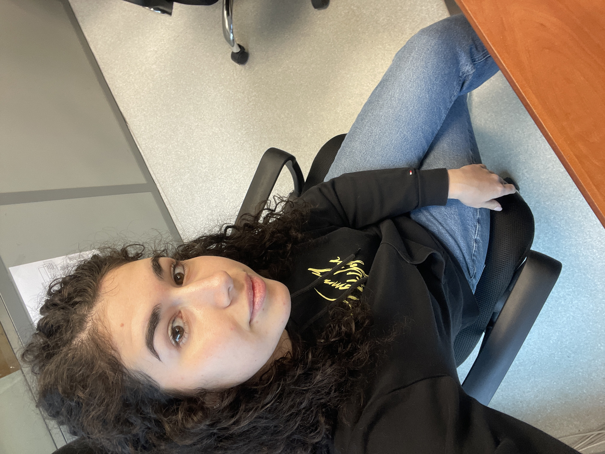 A young woman with curly dark hair, wearing a black hooded sweatshirt and light blue jeans, sits in a black office chair with a brown wooden desk to her right. She is looking up at the camera. The floor is light grey with dark grey or black accents. Part of an office chair with metal legs is visible in the upper center of the image. Part of a window or wall is visible in the upper left corner. The location in Sofia, Bulgaria cannot be determined from the provided information.