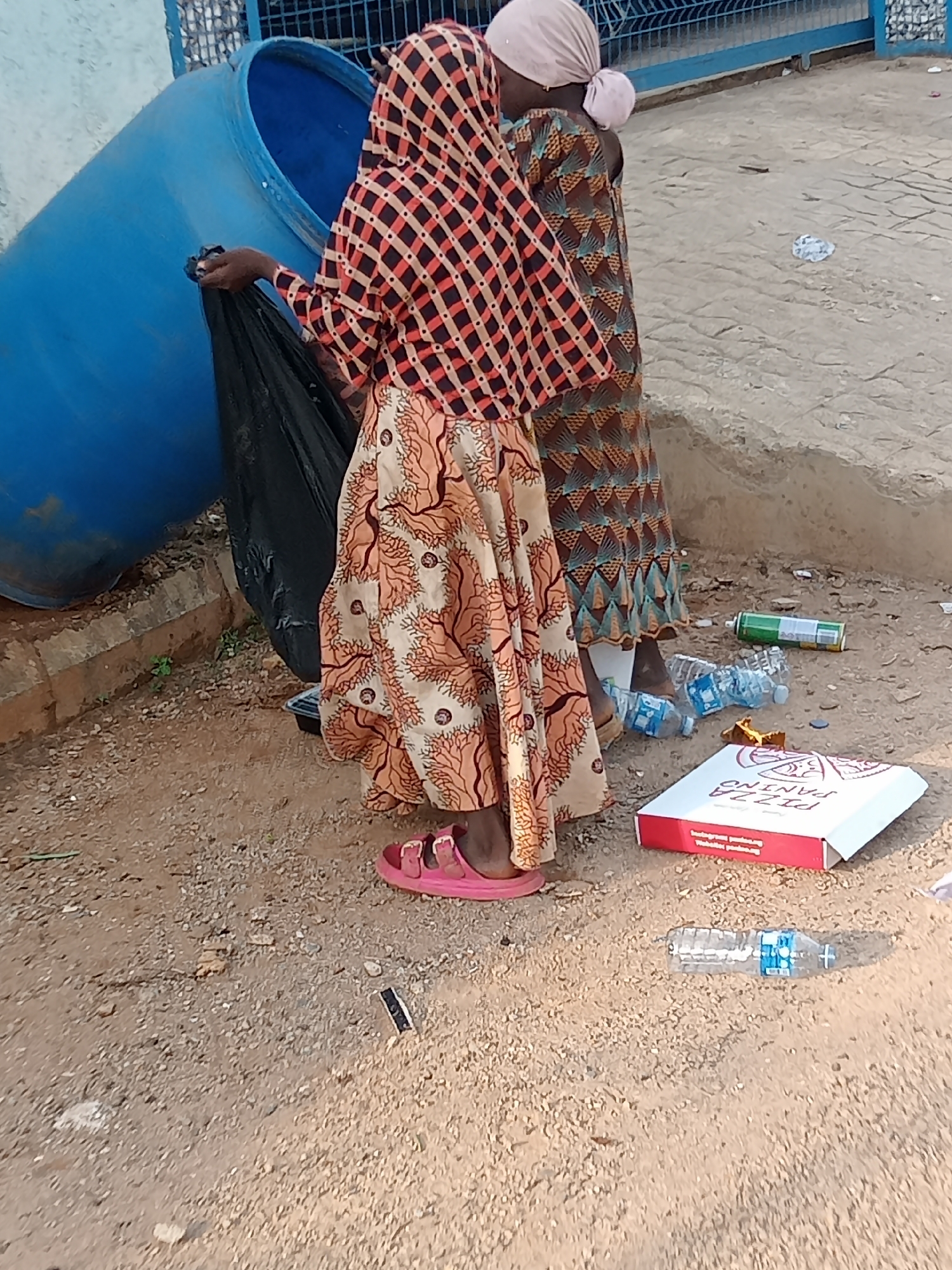 Two women are standing near a large blue trash container in Gwarinpa, Nigeria. One woman is placing a black trash bag into the container. The women are wearing patterned dresses and head coverings. The ground around them is littered with trash, including a pizza box, water bottles, and a spray paint can.