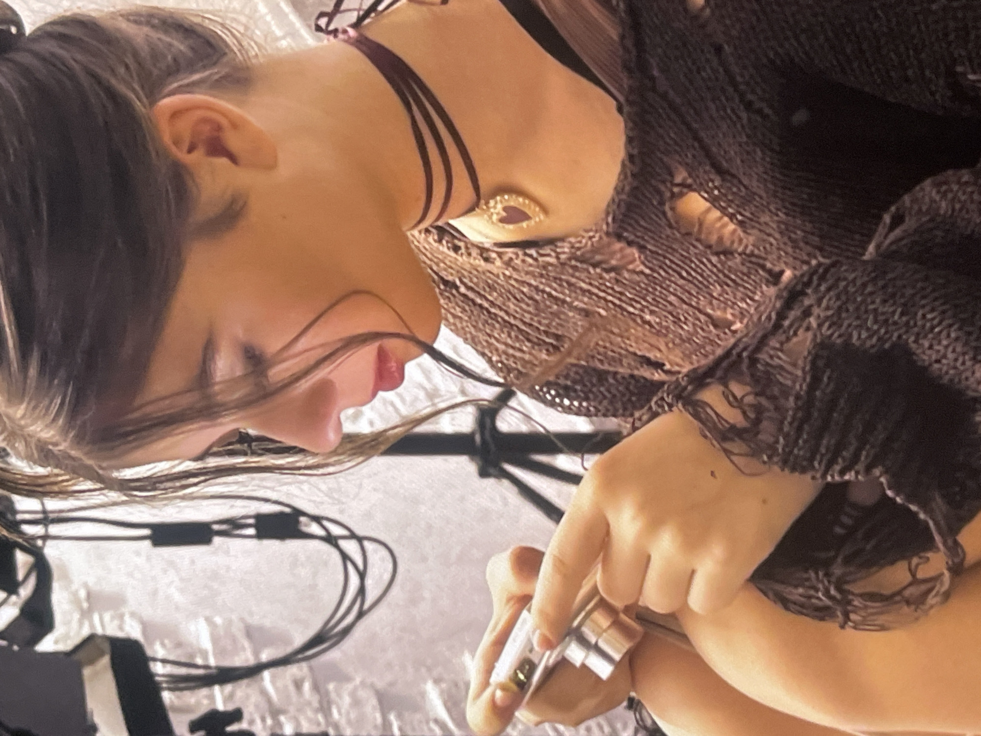 An eye-level shot captures a young person, likely female, in profile, looking downwards with a focused expression. Her dark brown hair is pulled back, with some strands falling forward around her face. She wears a dark, possibly distressed or ripped, knit sweater or top. Around her neck is a multi-strand dark choker, with a gold-colored pendant featuring a red heart attached to one of the strands on her chest.
Her hands are visible in the lower right, holding and manipulating a small, metallic, cylindrical object, which could be a vape pen, lighter, or small tool. Her fingers are wrapped around it, suggesting close attention to the item.
The background is indistinct but shows tangled dark cables, hinting at electronic equipment, against a textured, light-colored surface. A dark stand or structure is also visible behind the cables. The overall lighting appears to be artificial and internal, suggesting an indoor setting. The scene conveys a moment of quiet concentration. The activity takes place in Poltava, Ukraine. No text is visible in the image.