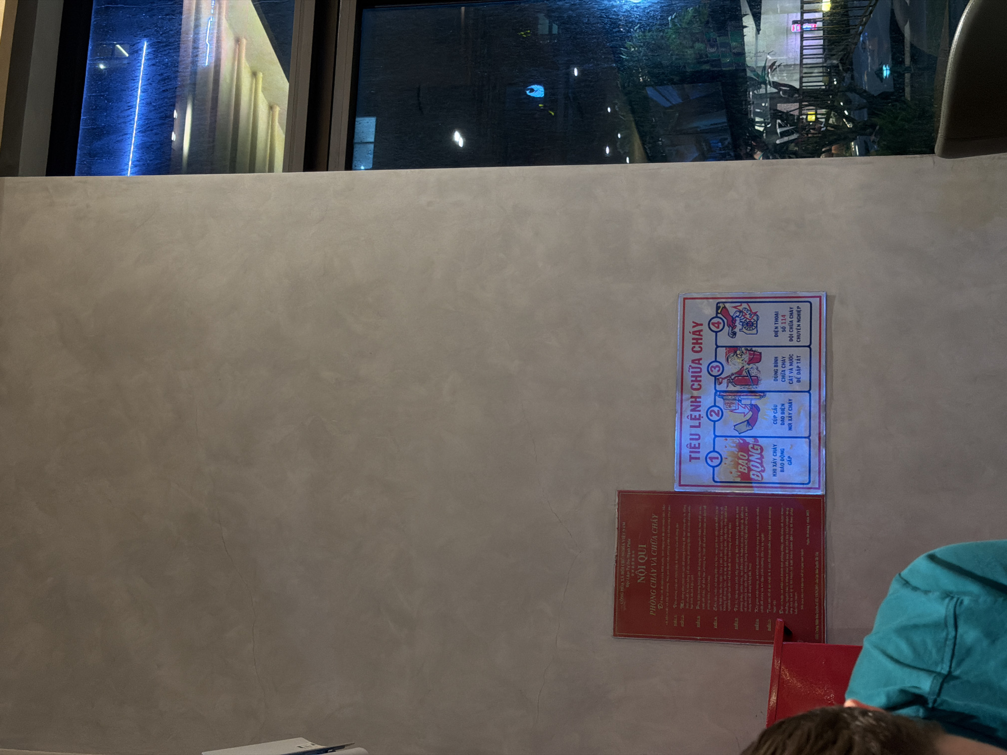 A man wearing a teal shirt is partially visible in the foreground of the image, his head and shoulders to the left. He appears to be seated and looking downwards. The background is dominated by a textured, light gray wall. Mounted on the wall are two red and white informational signs. The sign on the left, in Vietnamese, is titled "NỘI QUI PHÒNG CHÁY VÀ CHỮA CHÁY" (Rules for Fire Prevention and Fighting). It contains several numbered points of instruction. To the right of this sign is a smaller, rectangular sign with a blue border, also in Vietnamese, titled "TIỂU LỆNH CHỮA CHÁY" (Fire Fighting Procedure). This sign displays four numbered illustrations depicting steps for fire fighting, including using a fire extinguisher and calling for help. The right side of the image features a large window with a view of an exterior at night. Raindrops are visible on the windowpane, and the reflection shows blurred lights, buildings, and some vegetation. The scene suggests an indoor location, possibly a restaurant or public building, in Da Nang, Vietnam, given the language on the signs.