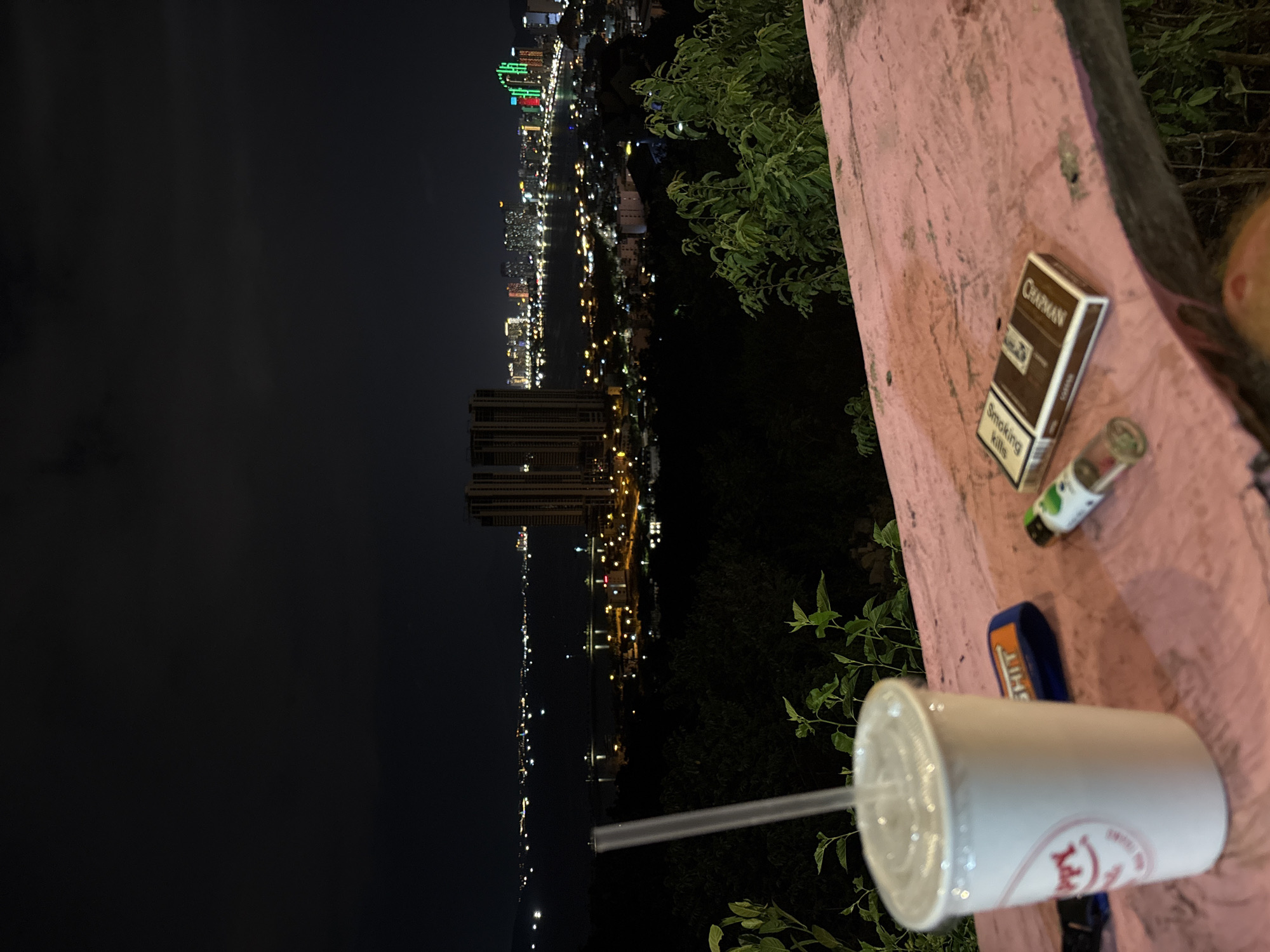 The image, taken in Bac Nha Trang Ward, Vietnam, shows a nighttime cityscape view from an elevated position. The city lights stretch along the coastline, with buildings and roads illuminated. In the foreground, a pink concrete ledge holds several items: a pack of Chapman cigarettes, a lighter, and a disposable cup with a straw. Vegetation is visible on the right side of the ledge. The sky is dark, contrasting with the bright city lights.