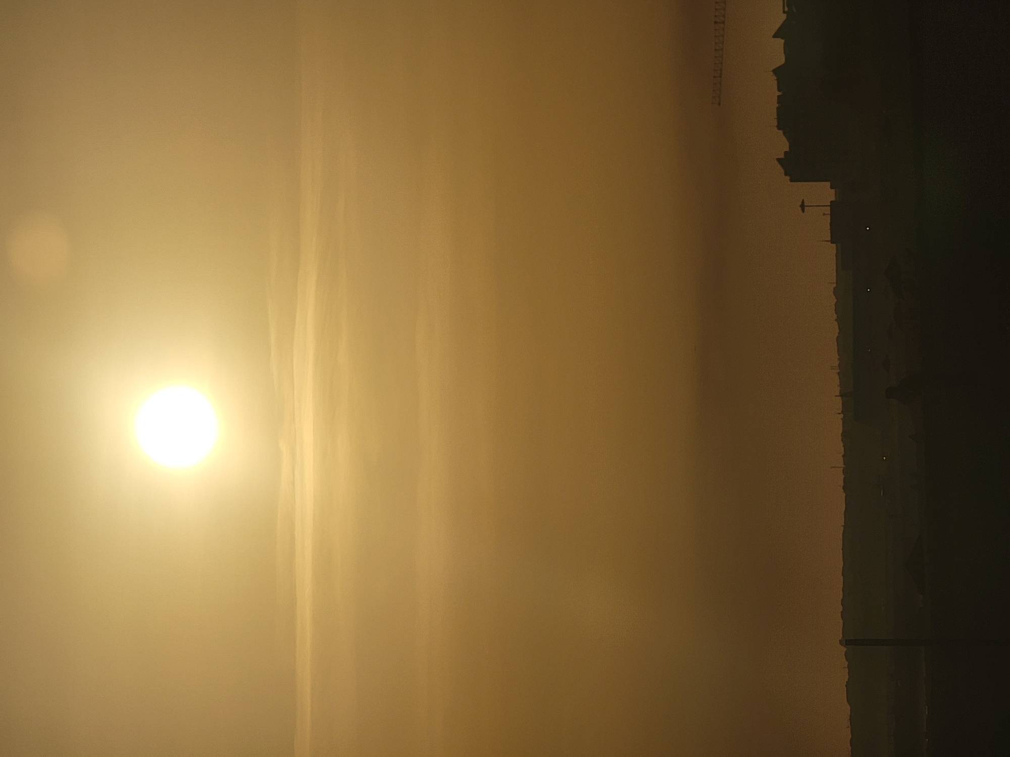 This media file depicts a hazy, possibly smoggy, sunrise over the coastal city of Quarteira, Portugal. The sun, a bright white orb with a halo of light, is positioned on the left side of the frame, its rays diffused by the atmospheric conditions. Streaks of lighter haze or clouds are visible against the golden sky. On the right side of the image, a dark silhouette of the Quarteira skyline emerges from the haze. This silhouette includes various buildings, some with distinct architectural features, and a prominent tall structure that appears to be a crane, suggesting ongoing construction or industrial activity. The overall scene conveys a sense of quiet dawn, with the obscured sunlight creating a soft, dreamlike atmosphere. The limited visibility hints at either pollution or dense morning fog, typical of coastal environments. The time of day is clearly early morning, as indicated by the sunrise. No people or specific activities are discernible, and there is no visible text.