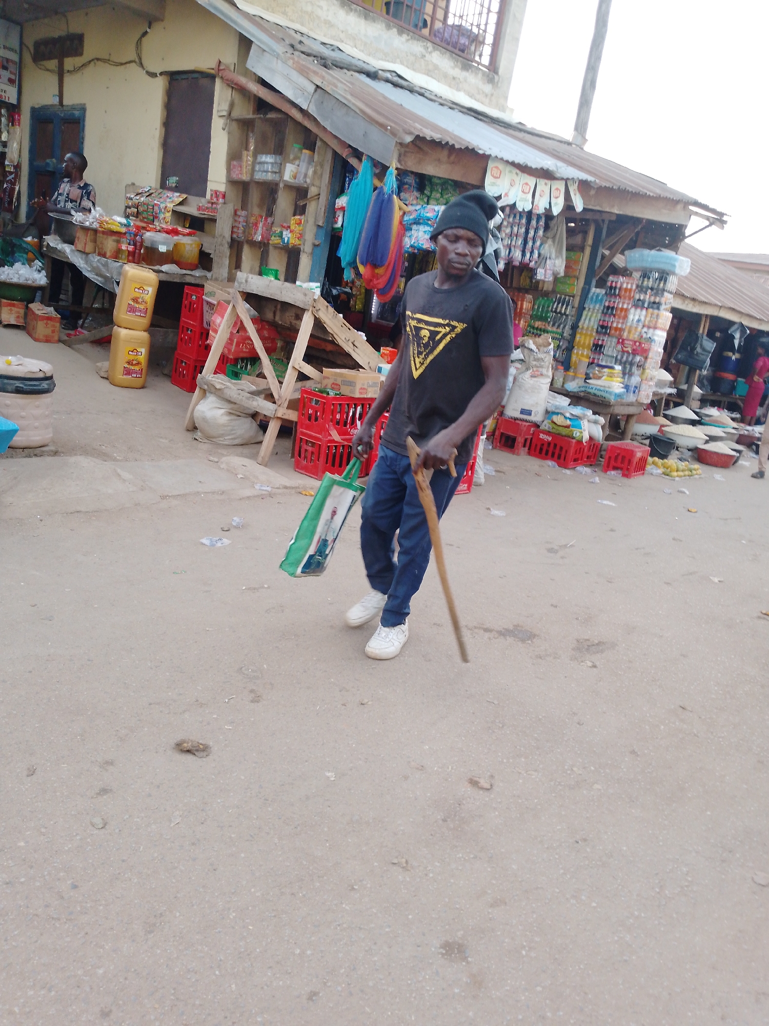 A man walks across a dirt and gravel street in an outdoor market scene in Akwanga, Nigeria. He is positioned in the center-left of the frame, moving towards the right. He wears a black beanie, a black t-shirt with a prominent yellow triangular graphic, blue trousers, and white sneakers. In his left hand, he carries a green and white reusable shopping bag, and in his right hand, he holds a wooden stick or cane. His expression is neutral.
Behind him and to both sides, numerous market stalls line the street. These stalls are constructed with wooden frames and covered with corrugated metal roofs. They display a variety of goods, including packaged snacks, bottled drinks, and large yellow plastic containers labeled "ROYAL OIL." Red plastic crates are stacked around the stalls, and sacks of what appear to be grains or produce are visible on the ground and on tables. On the far left, another person, possibly a vendor, is visible behind a stall laden with goods. The general ambiance suggests a busy daytime market. The sky appears bright but slightly overcast, and there are no strong shadows.