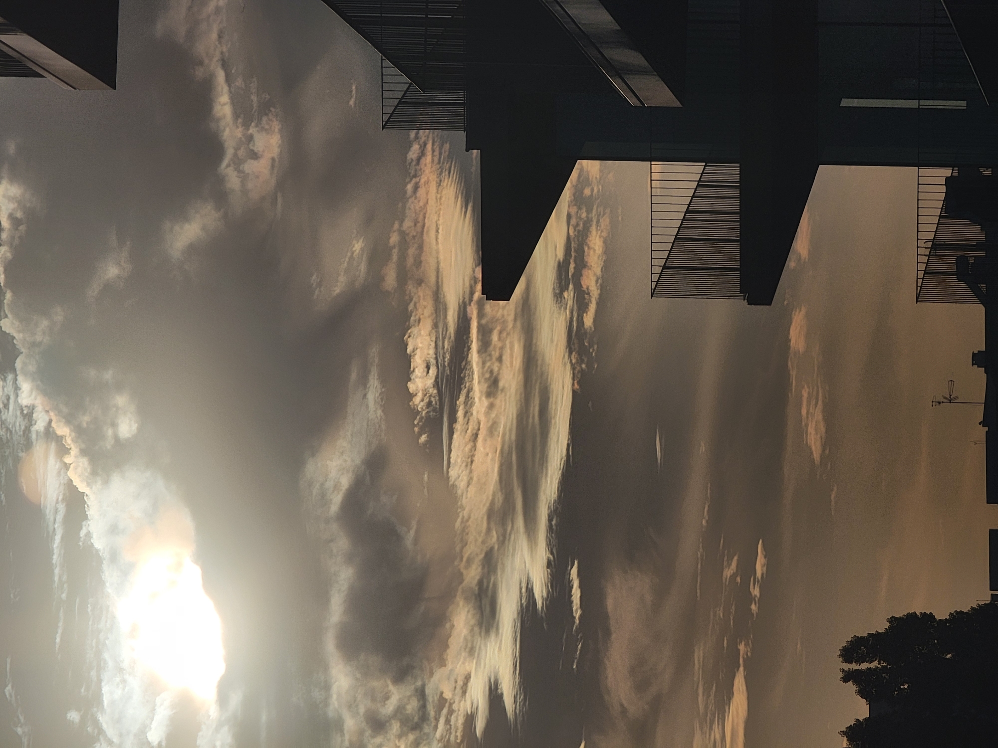 The image depicts a cloudy sky over the city of Quarteira, Portugal, at either dawn or dusk. The sun is visible behind a thick layer of clouds in the upper left quadrant, casting a bright, diffused light that gives the sky a warm, golden hue. The clouds are varied in texture and density, with some appearing wispy and others more solid and dark.
In the foreground and right side of the image, the dark silhouettes of a modern building's balconies are prominently featured. These balconies have metal railings and jut out from the building's structure, creating sharp geometric shapes against the sky. The building itself is not clearly visible beyond these architectural elements.
In the lower left corner, the dark silhouettes of trees are visible, suggesting a green space near the urban environment. The very bottom of the image shows the tops of other buildings, including what appears to be an antenna, indicating an urban setting. No people or specific actions are depicted. The overall mood is atmospheric and somewhat dramatic due to the lighting and cloud formations.
