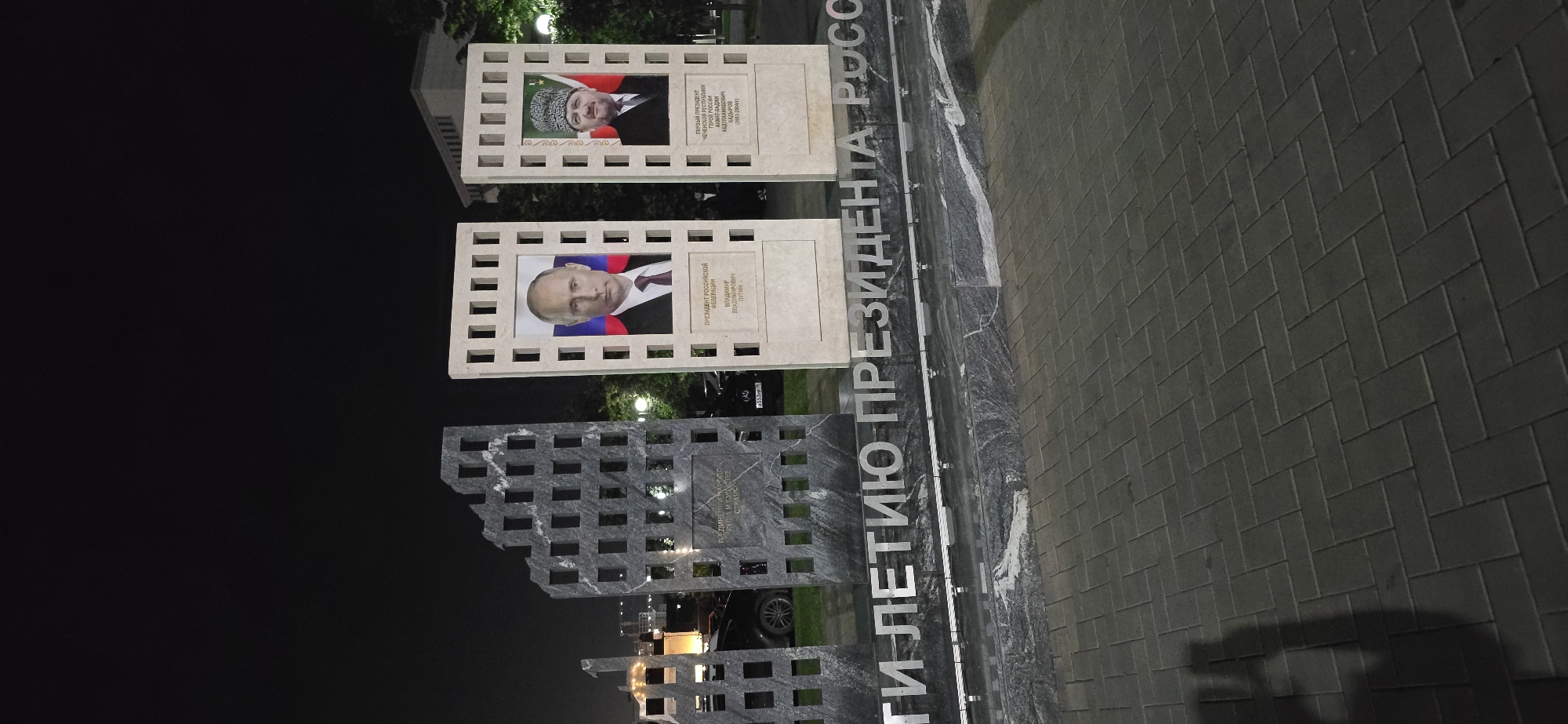 The image shows a monument, presumably in the city of Grozny, Russia, given the portraits included. The monument features portraits of Vladimir Putin, the President of the Russian Federation, and Akhmat Kadyrov, the first president of the Chechen Republic and a Hero of Russia. A gray granite structure is also present. The inscription on the monument reads: "To the Anniversary of the President of Russia." The monument is illuminated at night, and the scene also includes a paved area and some greenery in the background.