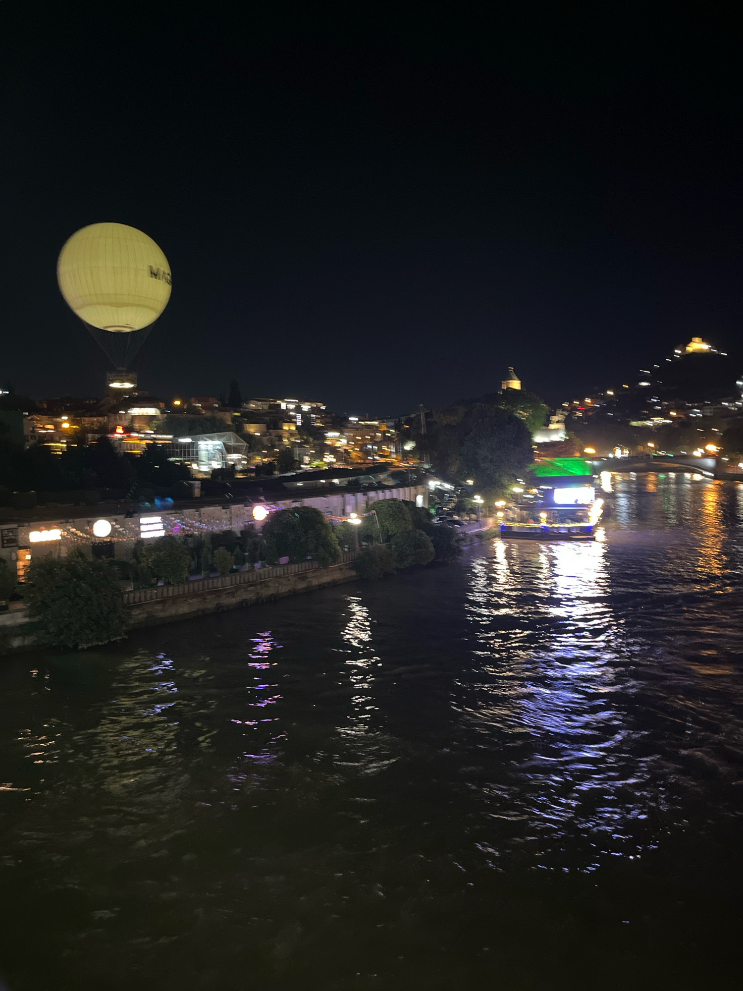 An illuminated hot air balloon hovers over the Mtkvari River at night in Tbilisi, Georgia. The balloon, a pale yellow, is emblazoned with the word "MAG" in dark lettering. Below, the river reflects the city's lights, casting shimmering patterns on the dark water. Along the riverbank, buildings are lit up, some with festive string lights. A boat with a green illuminated roof is docked on the river. In the distance, hills are visible, dotted with lights, suggesting a town or city spreading across the landscape. The sky is dark and clear, characteristic of a nighttime scene.