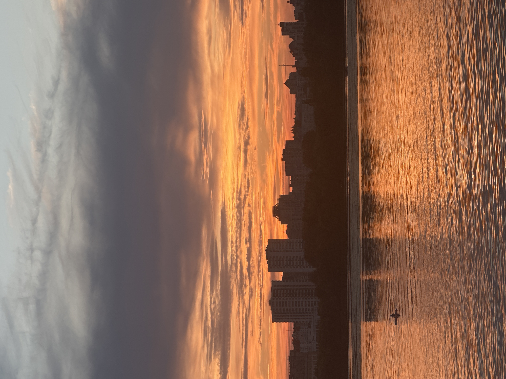 This image captures a serene sunset over the city of Lipetsk, Russia. The sky is a vibrant tapestry of orange and purple hues, with wispy clouds stretching across the horizon. Below the sky, a silhouette of the city's skyline is visible, featuring several tall buildings. In the foreground, a calm body of water reflects the colors of the sunset, creating a shimmering effect. A lone kayaker is present on the water, their small craft creating ripples that disturb the otherwise smooth reflection. The overall scene evokes a sense of peace and tranquility, with nature's beauty complementing the urban landscape.