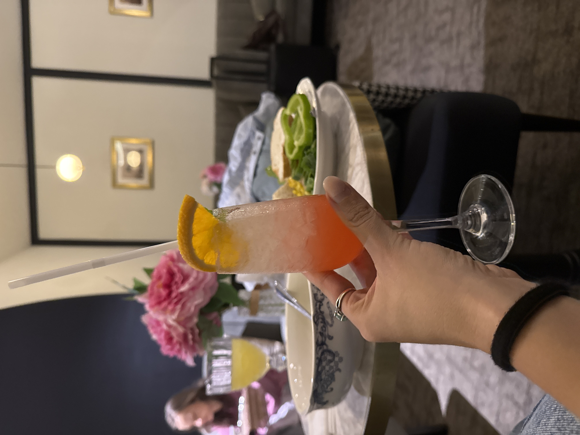 A close-up shot shows a hand holding a tall glass filled with an orange and white layered drink. The drink has a slice of orange on the rim and a white straw extending upwards. In the background, a blurred table holds a plate with salad, a partially visible blue and white bowl, and another smaller glass with a yellow drink. Pink flowers are also visible in the foreground. The setting appears to be an indoor space, possibly a restaurant or home, with framed pictures visible on the wall in the upper left corner. The lighting suggests it could be daytime or evening. The overall mood is relaxed and social.