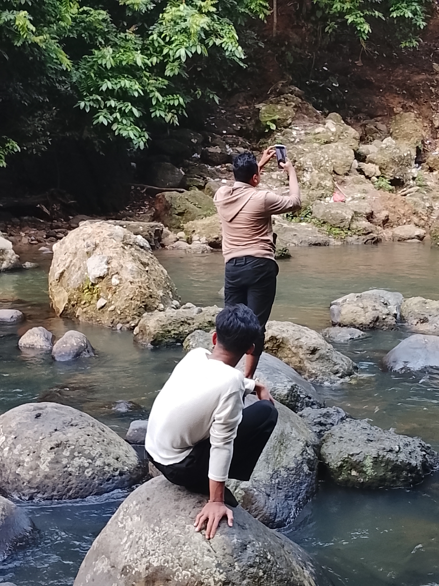 Two individuals are present in this outdoor scene. The person in the foreground is wearing a white long-sleeved shirt and black pants, and is crouching on a large rock in a river. The second person, in the background, is wearing a light brown hooded shirt and black pants and is standing on rocks, holding a mobile phone up to take a picture. The environment appears to be a natural setting with a river, large rocks, and lush green foliage in the background. The water is clear, revealing the rocky riverbed. The overall impression is that of a scenic, natural spot, potentially a tourist destination or a place for outdoor recreation. There is no specific indicator in the image that places this scene within Lhokseumawe, Indonesia, however, the overall landscape is consistent with many natural areas found throughout Indonesia.