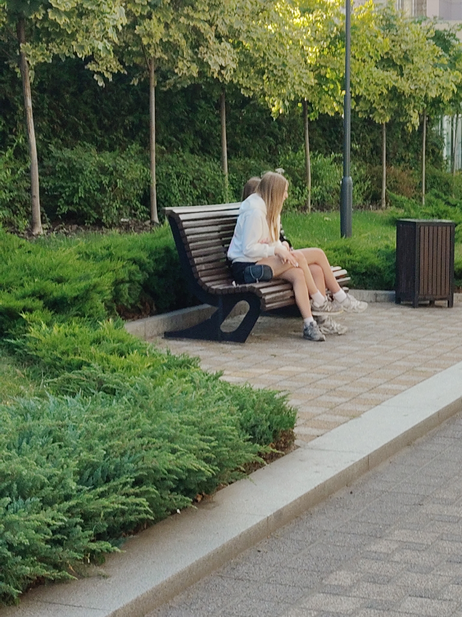 Two young women are sitting on a curved wooden bench in an outdoor park area. The woman on the left has blonde hair and is wearing a white hoodie and light-colored shorts. The woman on the right is mostly obscured by the first woman but appears to also have blonde hair and is wearing shorts and sneakers. They are sitting side-by-side, facing away from the camera, towards a line of trees and a building in the background. The bench is situated on a paved walkway with a textured stone pattern. To the right of the bench is a dark brown trash receptacle with a slatted design. The foreground of the image features lush green bushes. The setting is a public park with well-maintained landscaping.