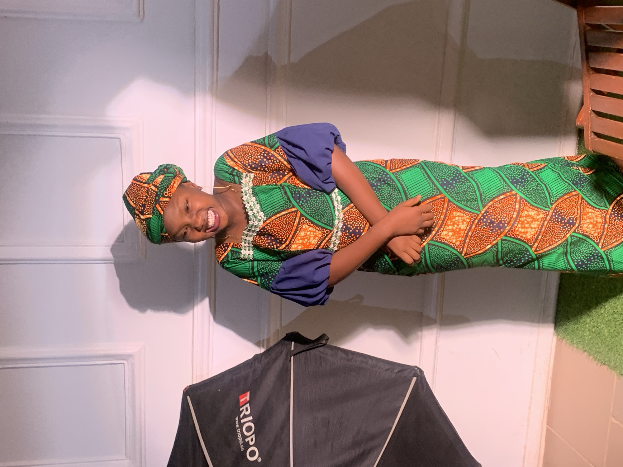 A woman smiles at the camera while wearing a green, orange, and blue dress with a matching head wrap. She is standing in front of a white door with a shadow on it. To the lower left of the frame is a black umbrella light. To the right of the door is a bit of green turf, a wood chair, and tan-colored tiles. The location is indoors in Barakin Akawo, Nigeria.