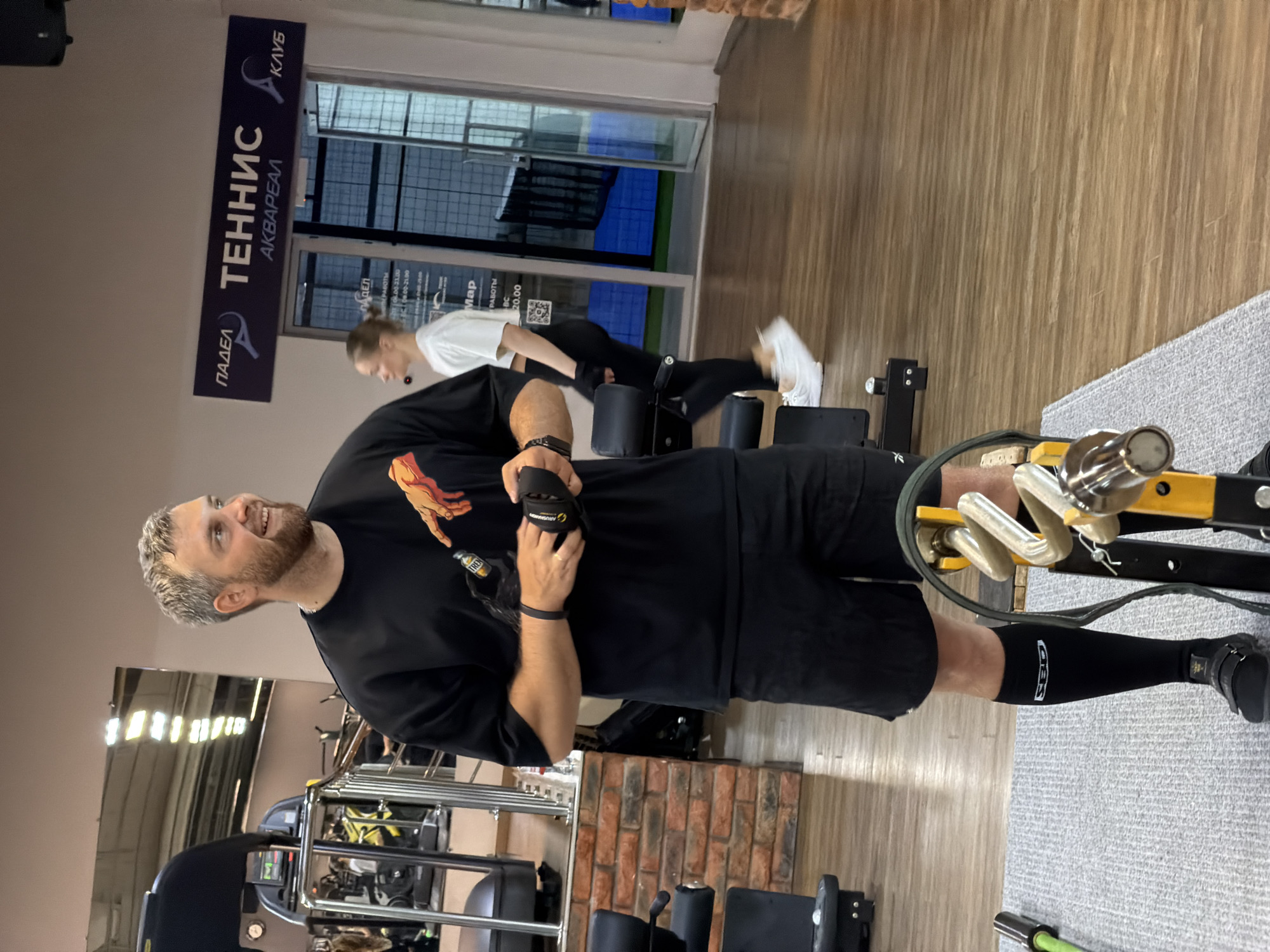 The image shows a man in a gym, likely in Chaikovsky, Russia, based on the "ПАДЕЛ ТЕННИС АКВАРЕАЛ КЛУБ" sign visible. The man is wearing a black T-shirt with a graphic on it, black shorts, black compression socks, and athletic shoes. He is holding what appears to be a weightlifting glove or wrist support. He is smiling and looking to his left. In the background, another person is exercising, performing a pushing motion. The gym equipment is visible, including a machine the man is interacting with, and general fitness equipment. The floor is a mix of grey carpet and wood. The sign "ПАДЕЛ ТЕННИС АКВАРЕАЛ КЛУБ" is mounted near a window, and through the window, a blue surface, possibly an athletic court or pool, can be seen. The interior walls are a mix of painted surfaces and brickwork.