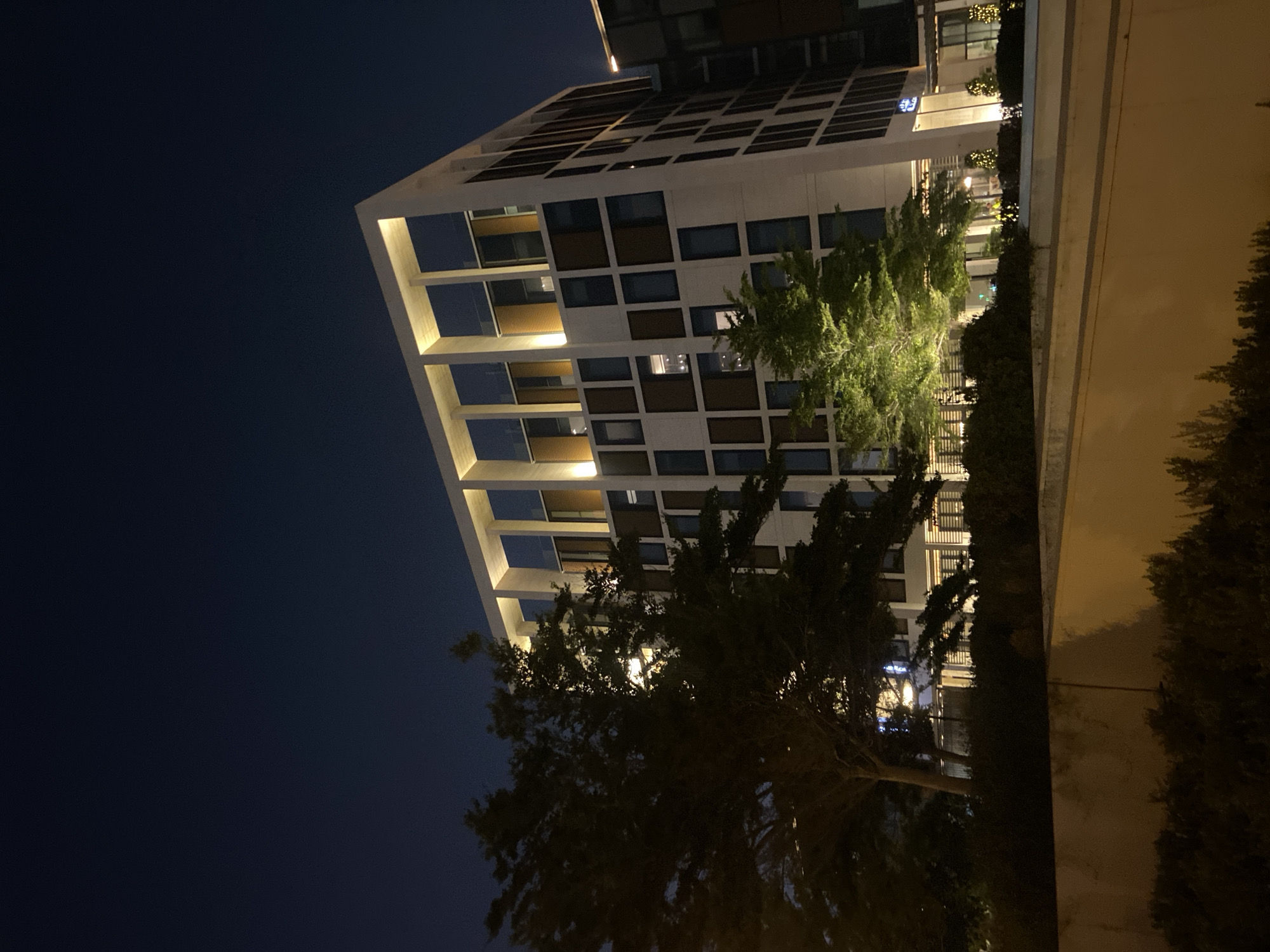 A nighttime exterior shot shows a modern, multi-story building in Deira, United Arab Emirates. The building features a white facade with a grid of windows and vertical architectural elements. Light spills from some of the windows, and the top floor is illuminated from above by recessed lighting. In the foreground, a concrete structure is visible, with some greenery and trees in front of the building, one of which is lit by a spotlight, casting a green glow. The sky is dark blue, indicating it is nighttime.