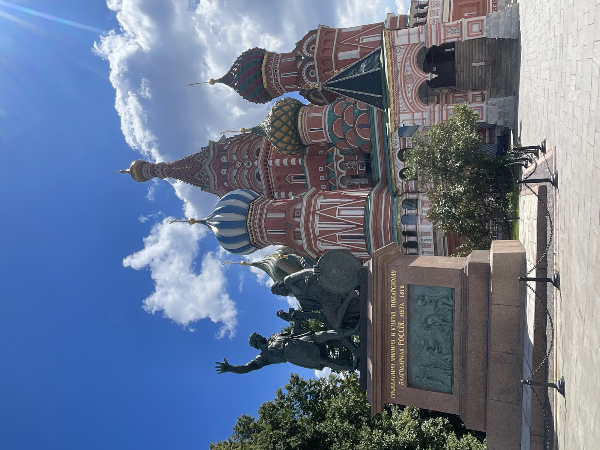 This is a vertical, eye-level photograph of the Monument to Minin and Pozharsky in front of Saint Basil's Cathedral on Red Square in Moscow, Russia. The sky is bright blue with scattered white clouds, suggesting a sunny day. The sun is casting light and shadows, indicating it is likely daytime.
The central subjects are the bronze monument and the cathedral. The monument depicts two men, Kuzma Minin and Dmitry Pozharsky, in historical attire, seemingly in the midst of a decisive moment. Pozharsky, to the left, has his right arm extended with his palm open, while Minin, on the right, holds a shield. The monument stands on a tiered stone pedestal. On the front of the pedestal, there is a bas-relief depicting a scene of battle or assembly, and above it, golden Cyrillic text. The text translates to: "TO THE CITIZEN MININ AND TO PRINCE POZHARSKY FROM A GRATEFUL RUSSIA. THE YEAR 1818."
Behind the monument, Saint Basil's Cathedral dominates the background. Its iconic onion domes, painted in vibrant red, green, and blue, are partially visible against the sky. The intricate architectural details of the cathedral, with its patterned walls and white trim, are also apparent. A paved walkway with a decorative chain barrier runs along the right side of the monument. Lush green trees are visible in the lower part of the frame, framing the monument and the cathedral. The overall atmosphere is one of historical significance and grandeur.