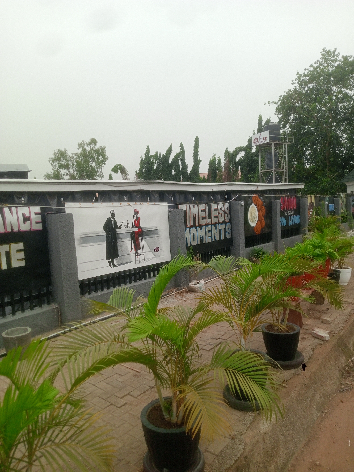 The image shows the exterior of a business in Gwarinpa, Nigeria. A fence separates the sidewalk from the property. The fence has several black panels. The first panel displays part of the word "ANCE," followed by the word "TE" on the second panel. The third panel features a black-and-white illustration of a man in a traditional Nigerian outfit standing next to a woman in a red dress seated at a bar. The next panel reads "TIMELESS MOMENTS" in gray lettering. The next panel has an image of a dish. The final panel displays the words "SAKOD" in a stylized font and "THE ART OF GOOD LIVING" below it. A water tank stands tall behind the fence. Potted plants line the sidewalk in front of the fence. The sky is overcast.