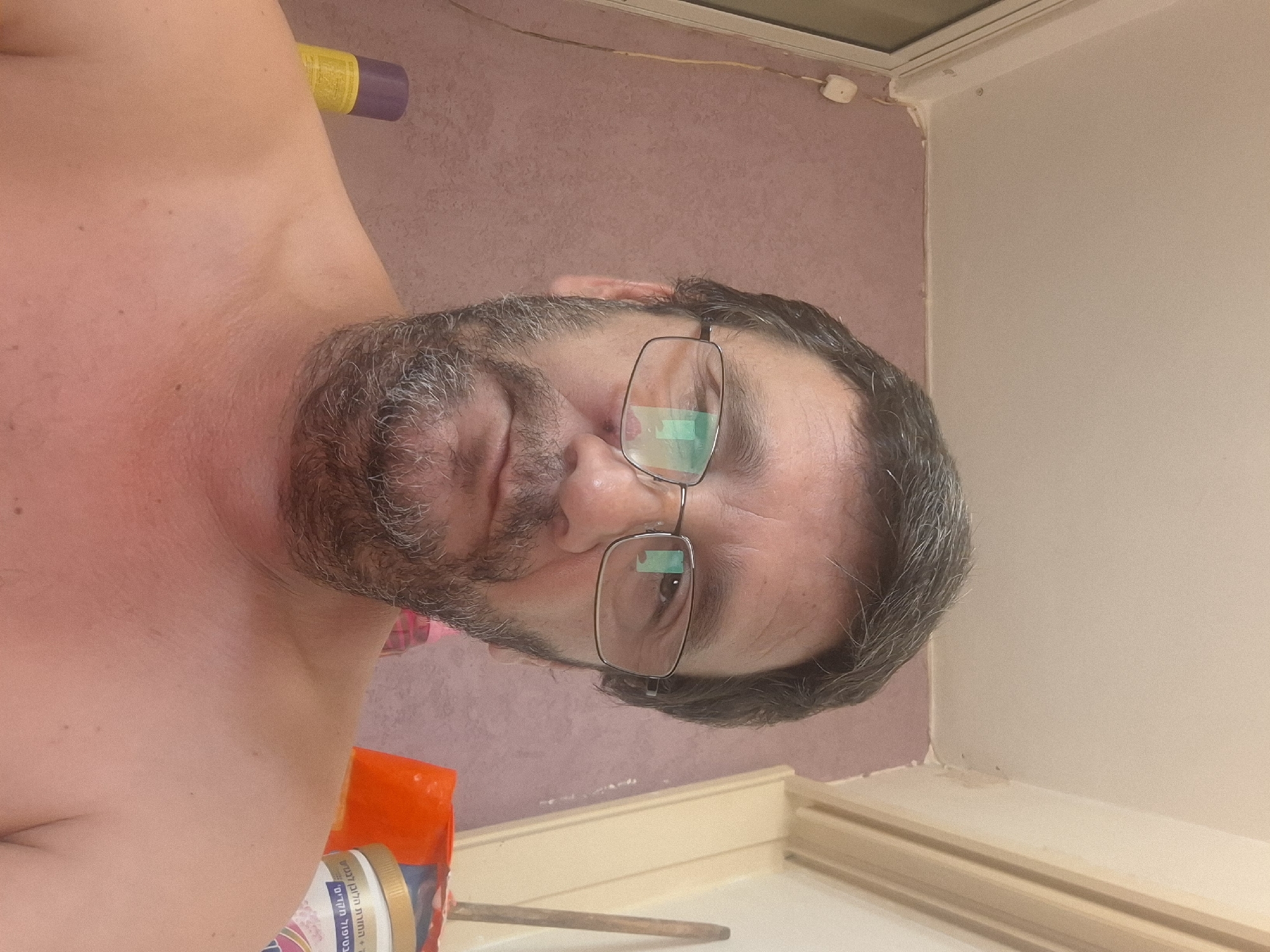 The image is a close-up selfie of a middle-aged man with graying dark hair, a beard, and glasses. He is shirtless, and his skin appears flushed, possibly from sunburn. The background shows a pink textured wall, a white electrical outlet with a yellow wire, and part of a ceiling. To the right, there's a glimpse of an orange object and a container with Hebrew writing that appears to be a skincare product. The scene is indoors, likely in a domestic setting. Without further information, the specific location in Yerocham, Israel, cannot be determined from the image alone.