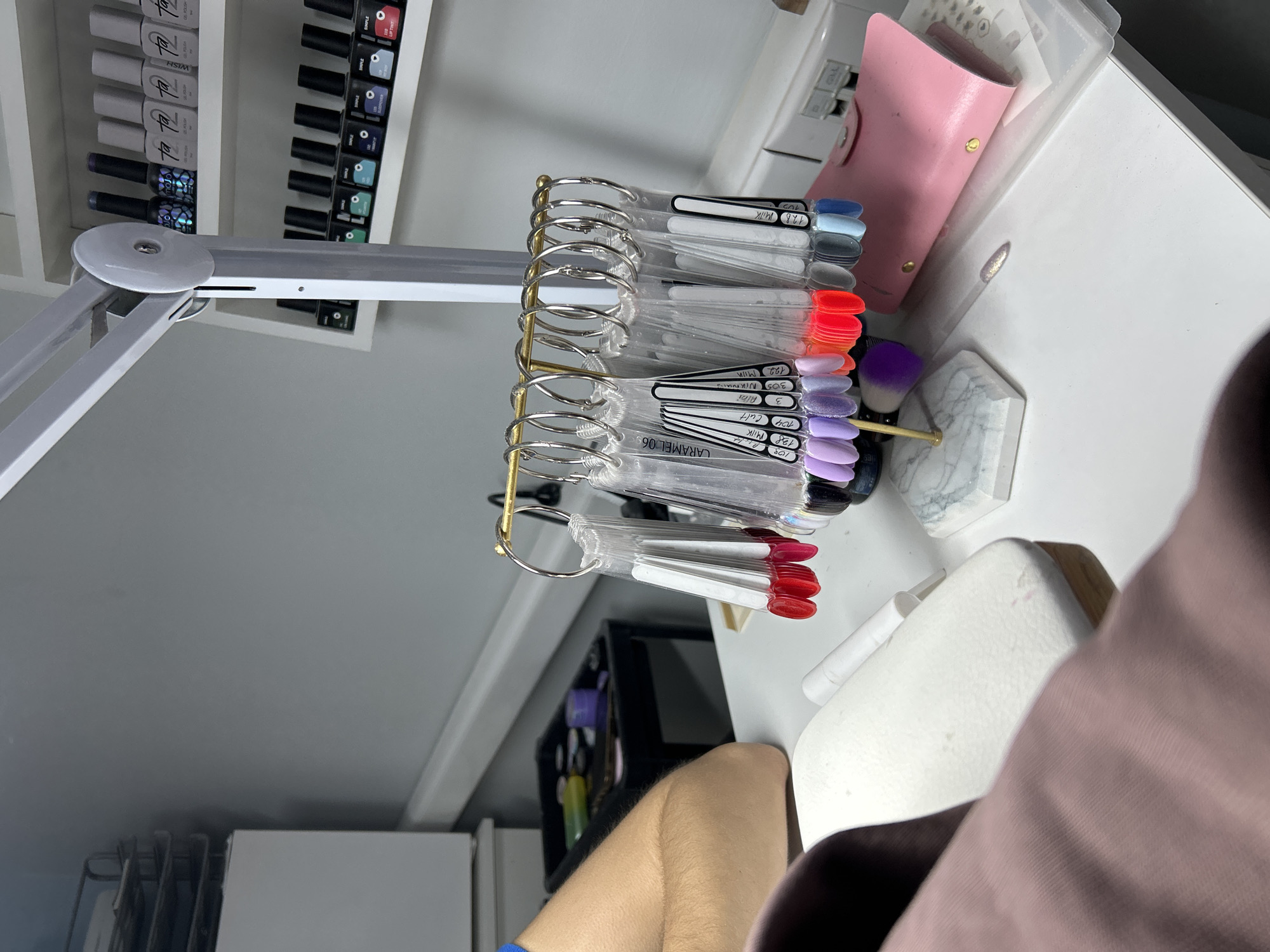 The image presents a beauty or cosmetic setup, dominated by a selection of nail polish color samples organized on a metal stand. These samples exhibit a range of colors including shades of blue, grey, red, orange, and purple, each labeled with identifiers such as "MILK" and "CARAMEL 06" along with numerical codes. Adjacent to the nail samples is a pink cosmetic case, a brush with a purple head, and a small, marble-patterned object. The background features shelves filled with nail polish bottles, indicating a professional or dedicated cosmetic space. A white articulated lamp extends into the scene from the left, providing illumination. The lower right portion of the image reveals a person's arm and clothing, implying the perspective of someone seated at the workstation. The location is not identifiable from the image.
