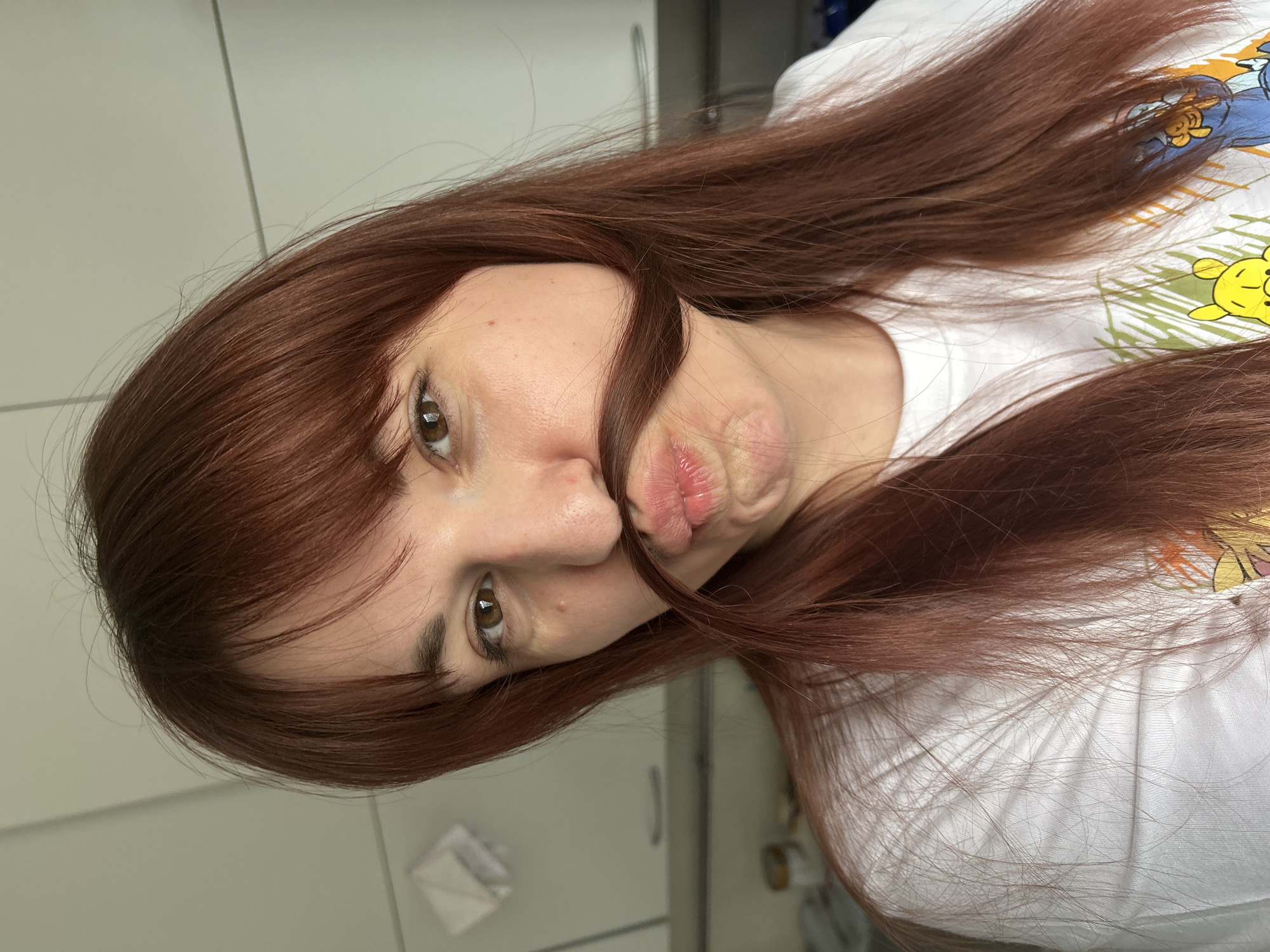 A woman with long auburn hair has pulled some of it down to resemble a mustache. She has light skin, brown eyes, and is wearing a white t-shirt with a cartoon print. The background is a light gray kitchen cabinet. The image does not indicate that it was taken in the city of Фоки, Russia.