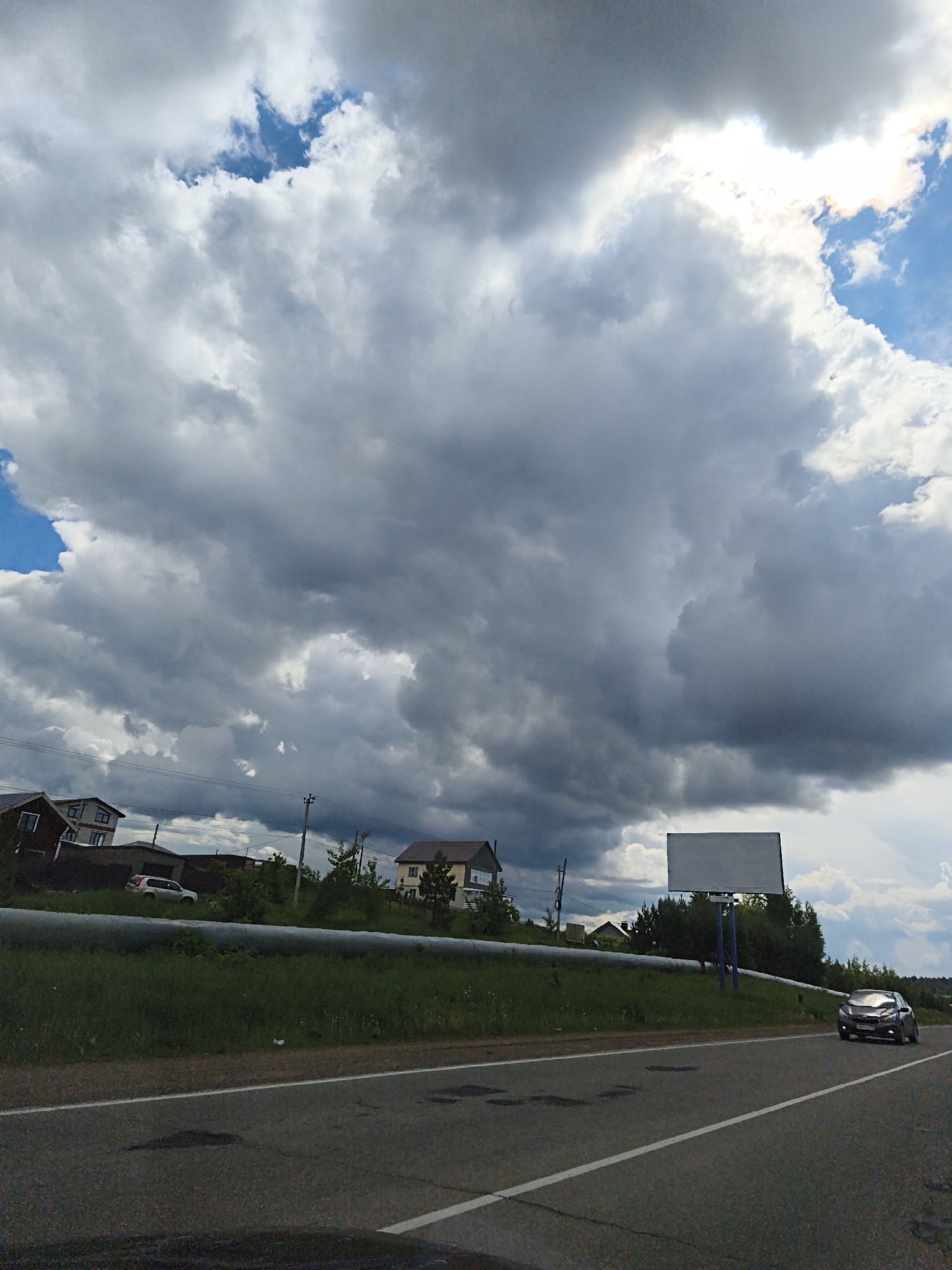 The image depicts a roadside scene in Прикамский, Russia, under a sky dominated by heavy, grey clouds with patches of blue visible. A two-lane asphalt road runs horizontally across the lower portion of the frame, with a car driving on the right lane. A green, grassy embankment runs along the roadside with a silver pipe running the length of the embankment. Behind the embankment, houses and trees are visible. A blank billboard stands on the embankment. Utility poles with wires run above the houses.