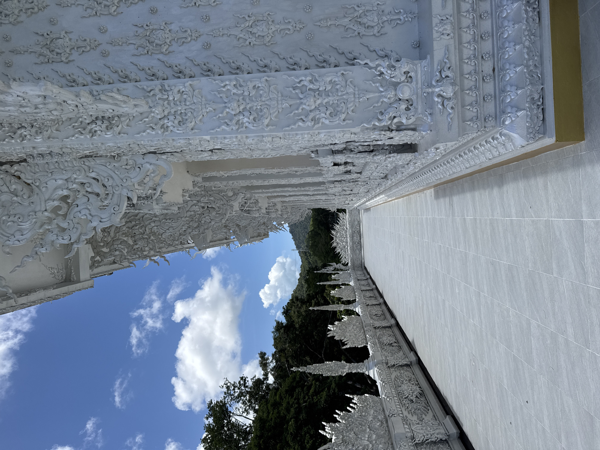 This is a side view of a white temple building with ornate, intricate white stucco decorations along the exterior walls and a balustrade. The building has a tiled walkway in front of it, which extends into the distance. The sky is blue with scattered white clouds. In the background, there are green trees and hills. The image appears to be taken during the day. The location is identified as Thep Krasatti, Thailand.