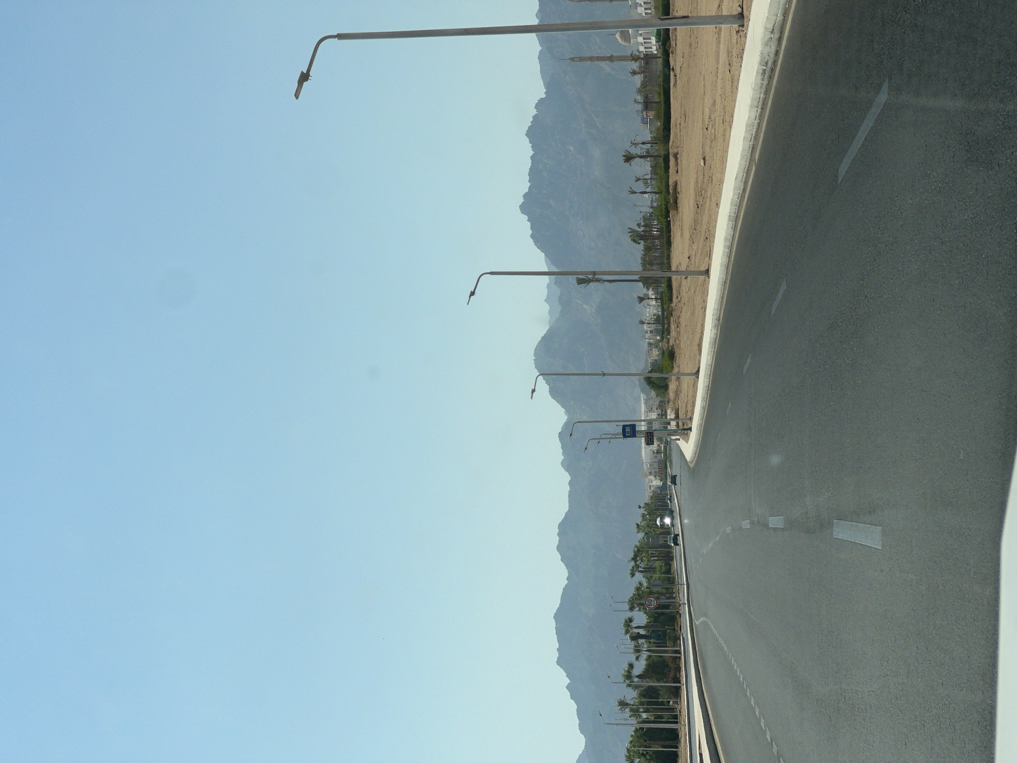 The image depicts a wide, paved road curving to the right, flanked by a sandy verge and palm trees. In the distance, a range of rugged mountains under a clear blue sky forms the backdrop. Several modern streetlights line the road. Buildings with white facades and a minaret are visible on the right side, suggesting a built-up area. Cars are present on the road, moving in the same direction. The overall scene is characteristic of a modern city or resort area, likely in a desert climate. Based on the context provided, the location is Sharm El Sheikh, Egypt.