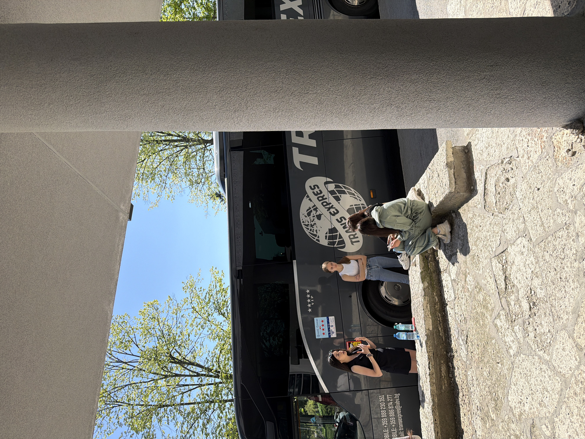The image presents an outdoor scene. A large, black bus, labeled "TRANS EXPRES" with its logo and contact details, dominates the frame's background. Adjacent to the bus, three young women are visible. One is kneeling while focused on something in her hands, another stands in a white tank top, and the third is in a black dress and appears to be using her phone. Water bottles sit beside the bus. The setting is partially framed by a stone structure, possibly a building's exterior. The backdrop features trees with light green foliage, suggesting spring or early summer, set against a blue sky. The location is Shumen, Bulgaria, based on the bus's phone number country code (+359).