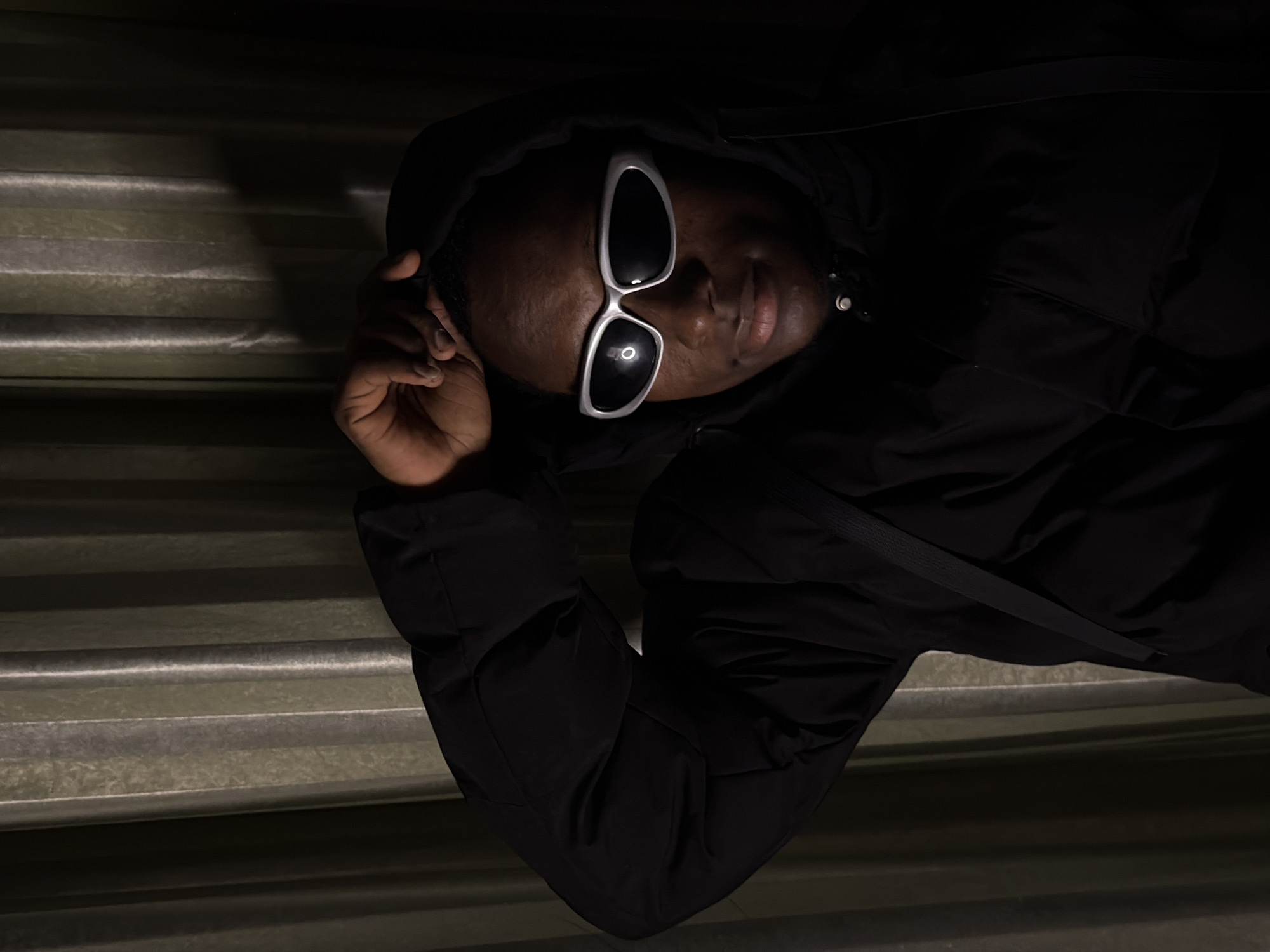 A person, wearing a black hooded jacket and futuristic silver-rimmed sunglasses, is positioned in front of vertical, textured curtains. The person's right hand is raised, touching the top of their head. The lighting creates a dramatic effect, with the upper portion of the image being brighter and the lower portion darker, casting shadows. The person's face is partially illuminated by the light source, highlighting their features. The background curtains are a muted green-grey color, with distinct vertical folds that catch the light. There is no discernible urban environment or specific location in Jiangjin, China.