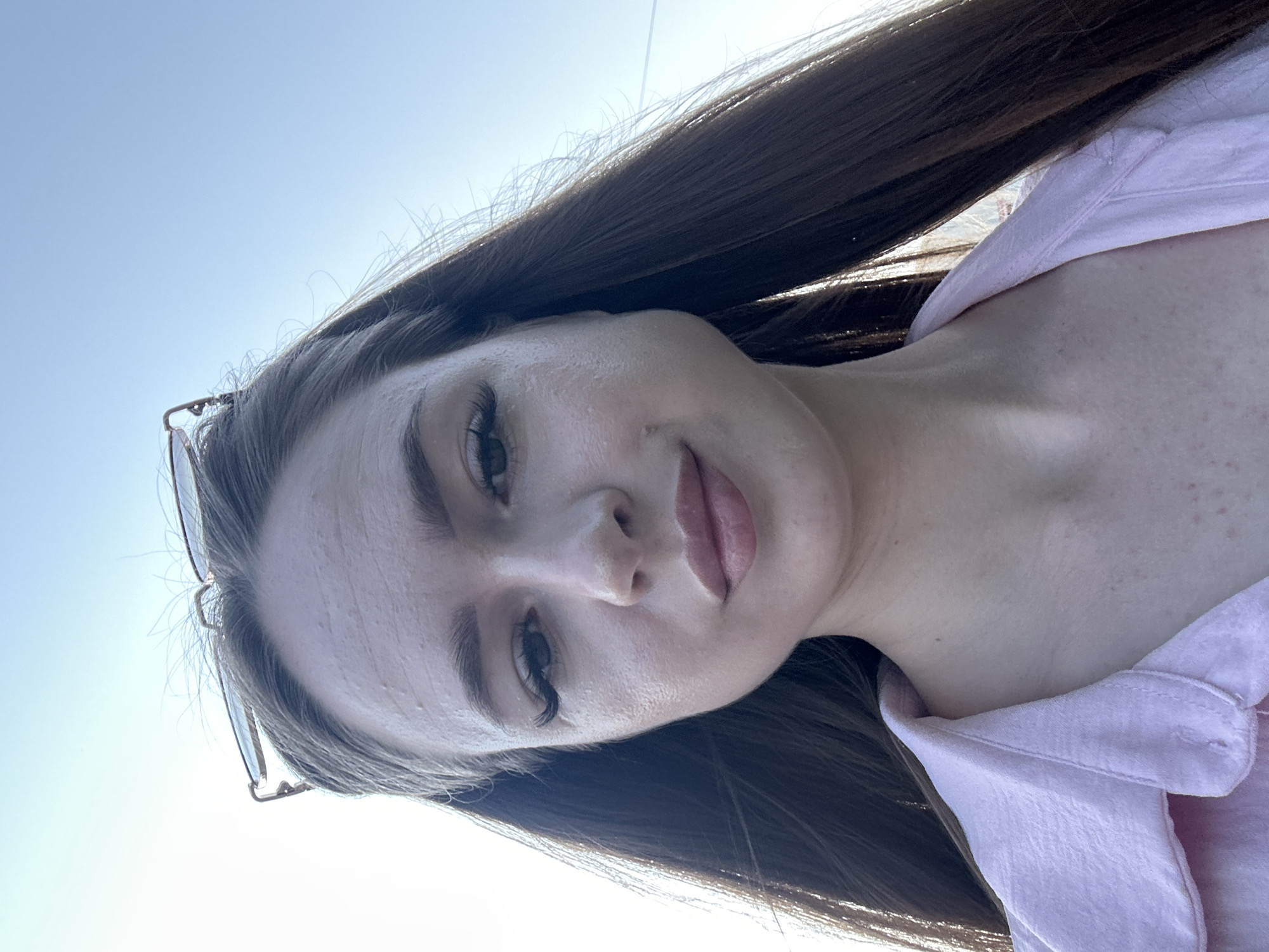 An outdoor, waist-up, selfie-style shot shows a fair-skinned young woman with long, dark brown hair that is parted in the middle and falls around her shoulders. She is looking at the camera and has on a light pink collared shirt. Her eyebrows are dark and well-defined, and she is wearing dark mascara on her eyelashes. She has on a light shade of lipstick that matches her shirt. Her forehead appears to have a few small raised bumps. She is wearing glasses on top of her head. The background is a clear, bright blue sky.