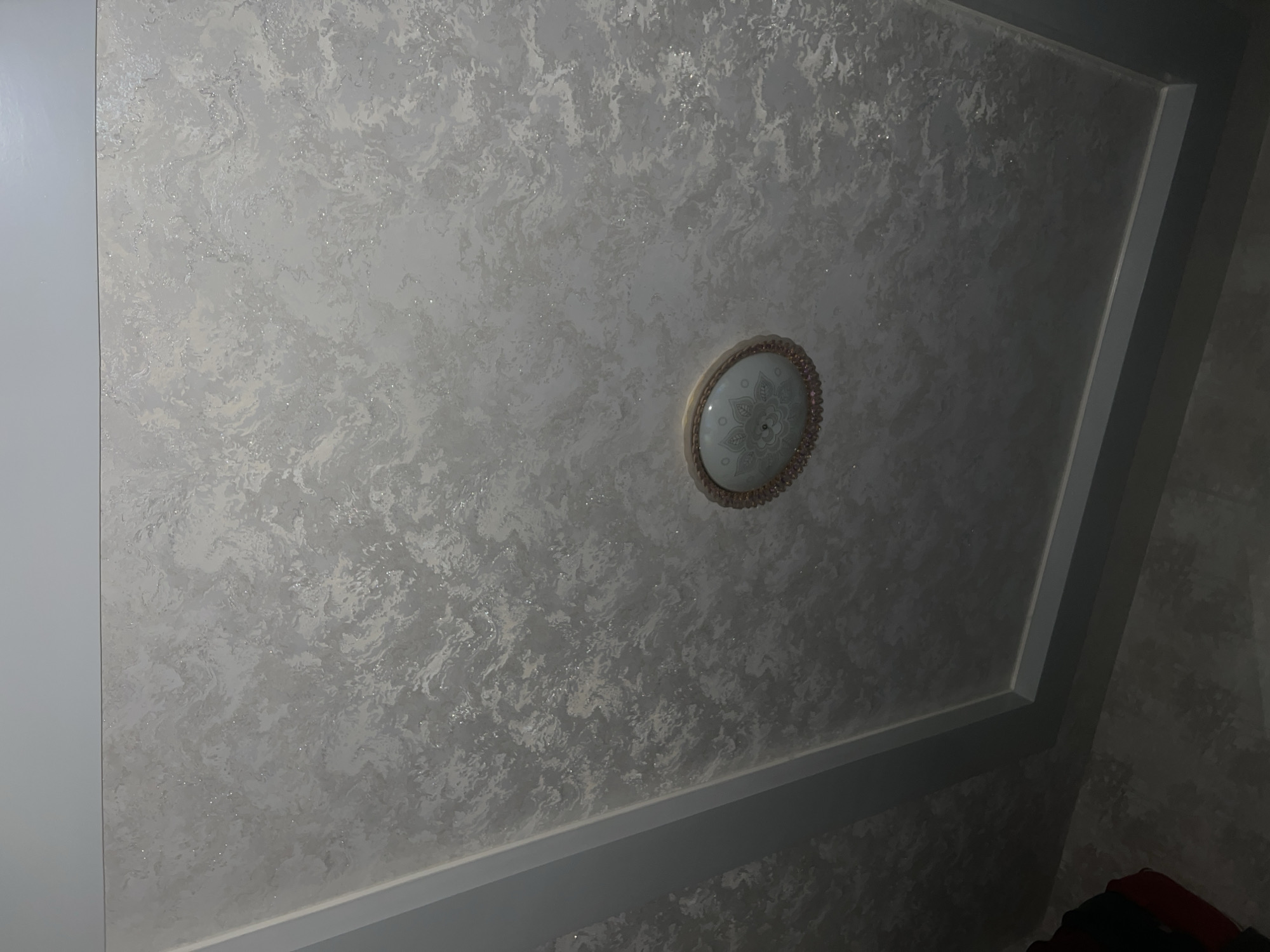 The image shows a close-up view of a textured ceiling with a decorative light fixture. The ceiling has a marbled or swirled pattern in shades of white and gray, with small, reflective flecks embedded within the texture, suggesting glitter or mica. The light fixture is oval-shaped and centrally located on the ceiling. It has a patterned glass cover and a beaded trim around its edge. The ceiling appears to be part of an interior space. The lower portion of the image shows a corner of a room where the wall meets the ceiling, with a simple, painted trim. A dark object, possibly furniture or clothing, is partially visible in the bottom left corner. The lighting is soft, illuminating the ceiling and the light fixture. No specific location in Yukari Chek, Uzbekistan, can be identified from this image.