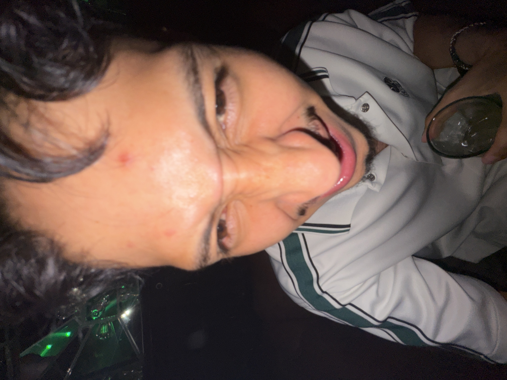 A close-up, rotated image depicts a person with dark hair, brown eyes, and tanned skin, making a distorted facial expression with an open mouth. The person wears a white collared shirt with green and black stripes along the collar and button line. A bracelet is visible on the wrist holding a glass filled with a clear liquid and ice. The background is dark and indistinct. The location is not discernible from the image. No information is available to place the image in San Bernardino Tlaxcalancingo, Mexico.