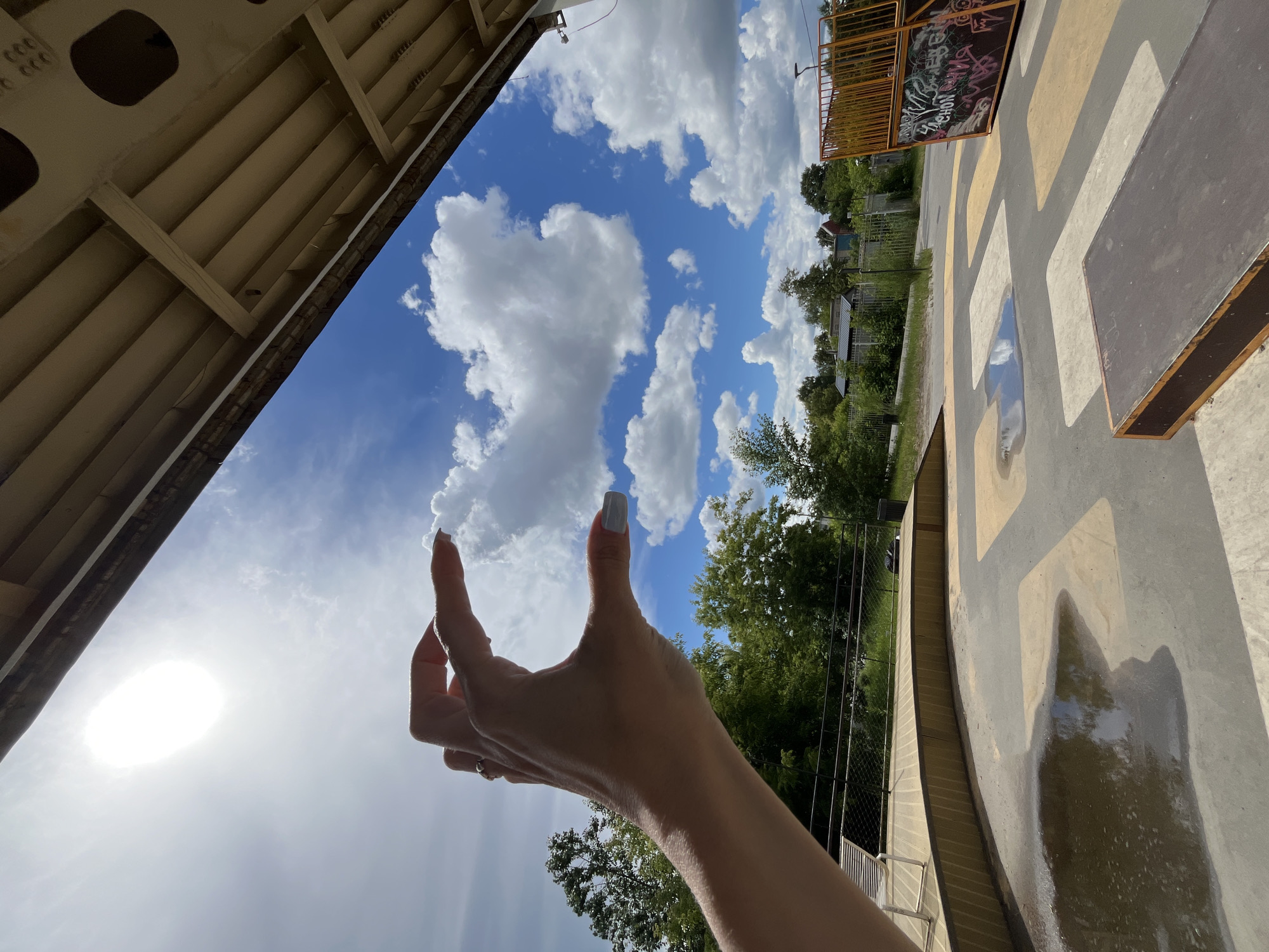 A person's hand, with fingernails painted a light gray, is held up to the sky, pinching a fluffy white cloud between their thumb and index finger. The sun is a bright, blown-out circle in the upper left quadrant of the image. Below the sky is a view of a skatepark, with wooden ramps, a concrete surface marked with yellow and gray shapes, and some puddles of water reflecting the sky. Trees and buildings are visible in the background. The skatepark is located at the Nizhny Novgorod Skatepark, located at ulitsa Vavilova, 1A, Nizhny Novgorod, Russia.