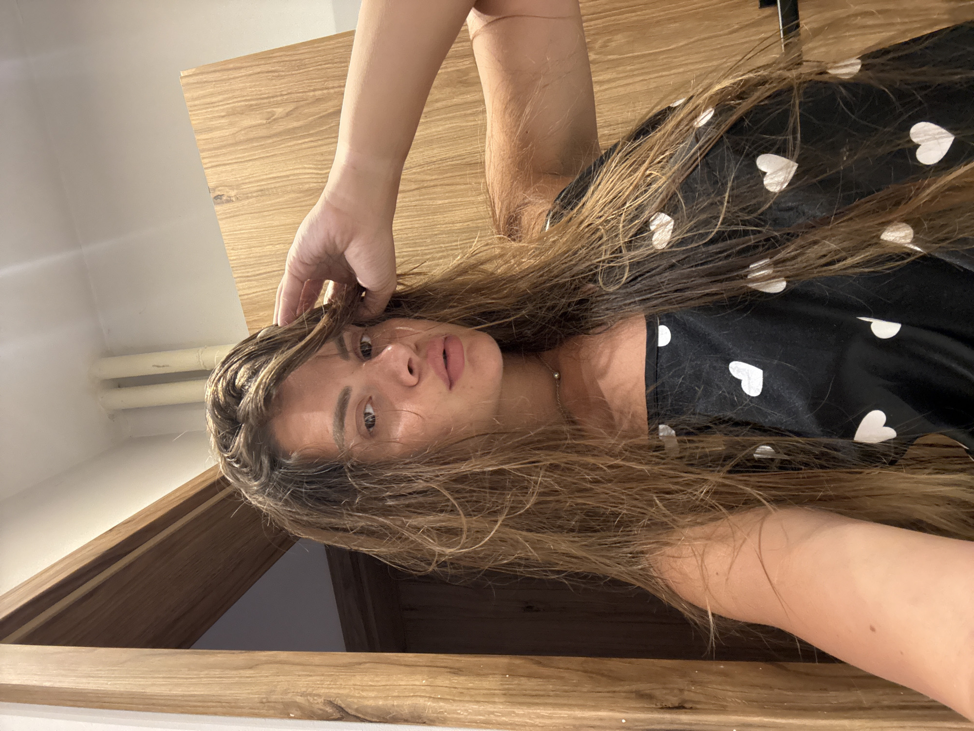 The image shows a woman with long, wet, brown hair and fair skin, wearing a black top with white hearts. She is looking at the camera and has her hand on her head. The background appears to be a room with wooden elements and white walls. There are no specific details in the image that would allow for identification of the location within the city of Plovdiv, Bulgaria.