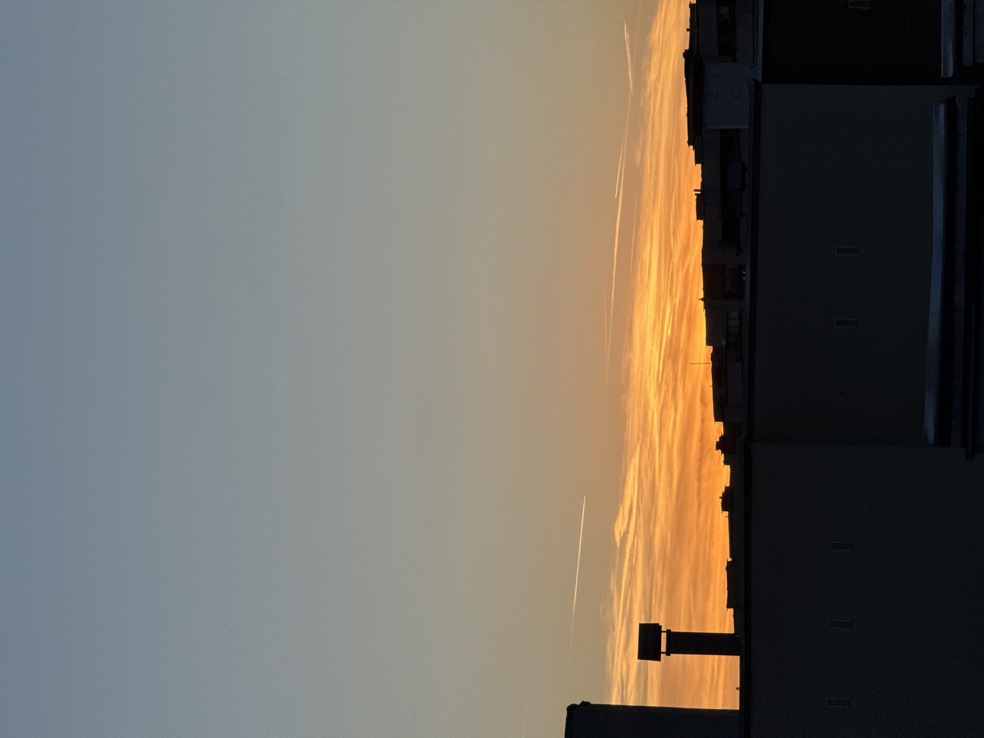 The image captures a sunset over the city of Pleven, Bulgaria. The sky transitions from a pale blue at the top to a vibrant orange near the horizon, with wispy clouds streaked across the sky. Two contrails from airplanes are visible. In the foreground, the dark silhouettes of buildings and a chimney are present. The buildings are modern, flat-roofed structures, suggesting an urban environment. The scene is devoid of any people or discernible actions. The time of day is clearly indicated by the sunset hues.