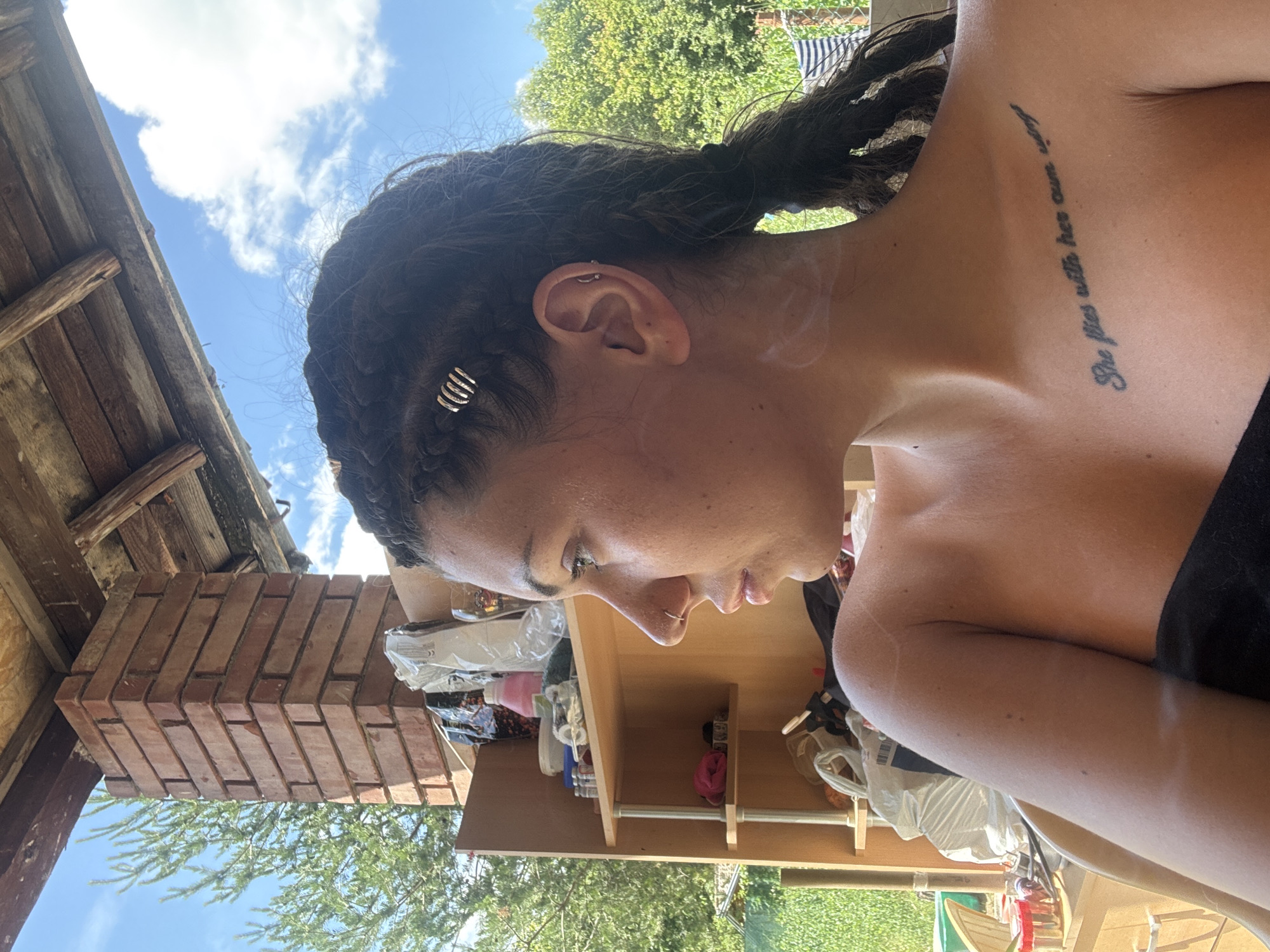In Belopoptsi, Bulgaria, a young woman with dark braided hair, accessorized with small silver rings, sits outdoors in what appears to be a covered patio area. She is facing to her right, with her head turned and her eyes looking to the side. Her left shoulder and upper chest are visible, and a tattoo on her chest reads "She flies with her own wings." The background shows a brick pillar, shelves with various items, green foliage, and a cloudy blue sky.