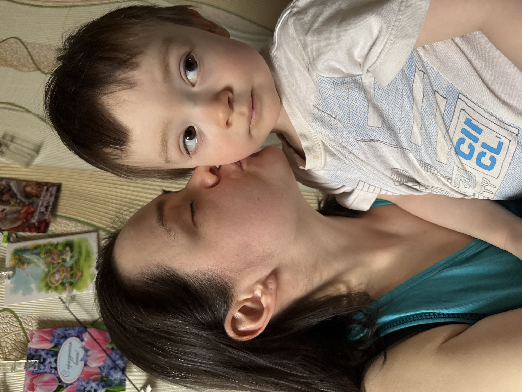 An intimate, close-up portrait captures a tender moment between a woman and a young child, likely indoors in Izhevsk, Russia. The woman, with dark hair, is lying on her side, kissing the child's cheek. She is wearing a teal tank top. The child, with light skin and dark hair, is positioned above her, looking directly at the viewer with wide, brown eyes. The child wears a light-colored t-shirt with blue and white graphic elements, including partially visible text that reads "CII CL". They are situated on a light-colored, subtly striped surface, possibly a bed or floor. Near the woman's head, several decorative cards or pictures are visible. One prominent card features pink tulips and blue hyacinths, bearing the Cyrillic inscription "С 8 Марта! Дорогая!", which translates to "Happy March 8th! Dear!". The image conveys a strong sense of affection and warmth between the subjects.