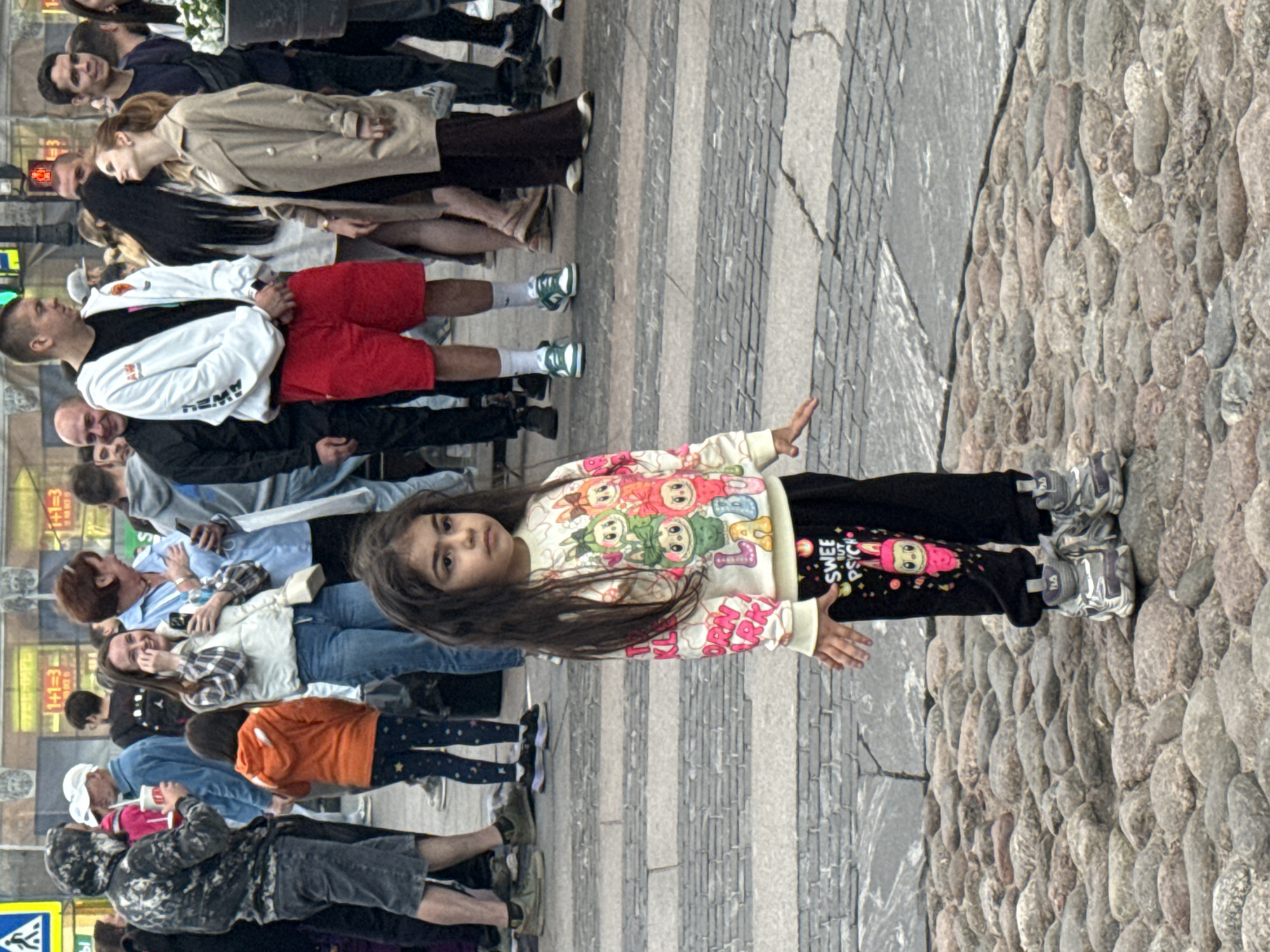 A young girl stands in the foreground on a cobblestone street, looking to her right with a neutral expression. She has long, dark, wavy hair and is wearing a white sweatshirt with colorful cartoon characters and black pants with a pink cartoon character and text that reads "SWEET BUT PSYCH." She has on gray sneakers with white laces. Behind her, a crowd of people are walking and standing. The background appears to be a city street with buildings and shops. The scene is likely a public square or pedestrian area in Saint Petersburg, Russia, given the architectural style and clothing of the people. The cobblestone paving and the presence of multiple individuals suggest a busy urban environment.